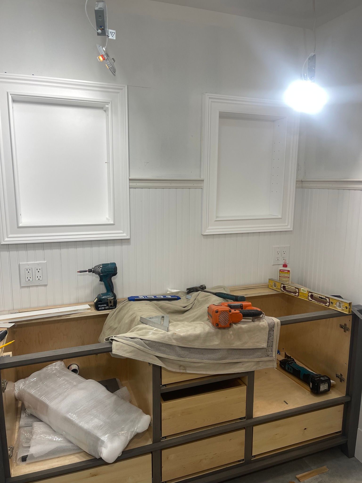 A vanity cabinet base being installed in a bathroom with white wainscoting and two framed wall mirrors.