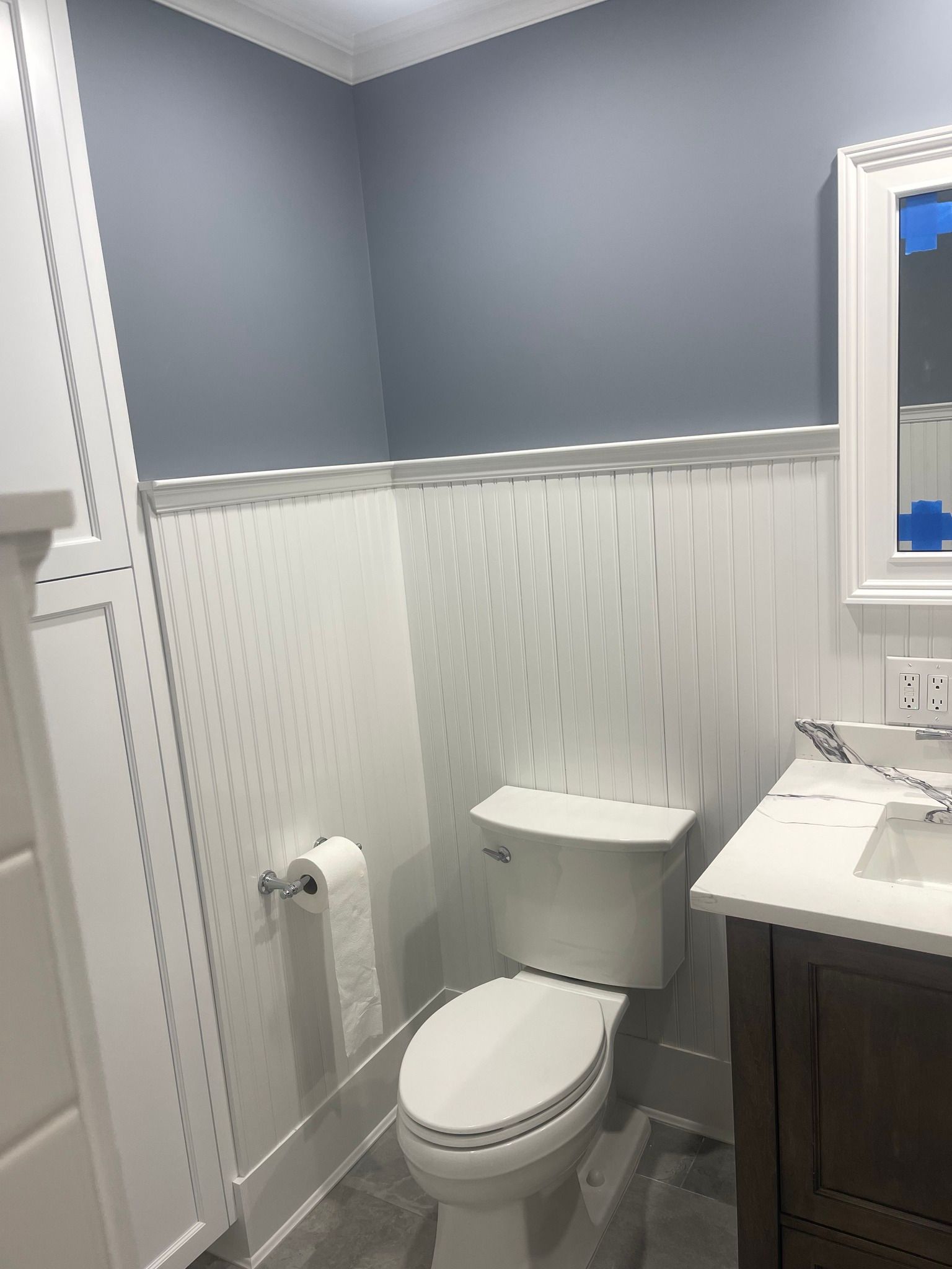 A bathroom featuring blue walls, white beadboard wainscoting, a white toilet, and a vanity with a white countertop.