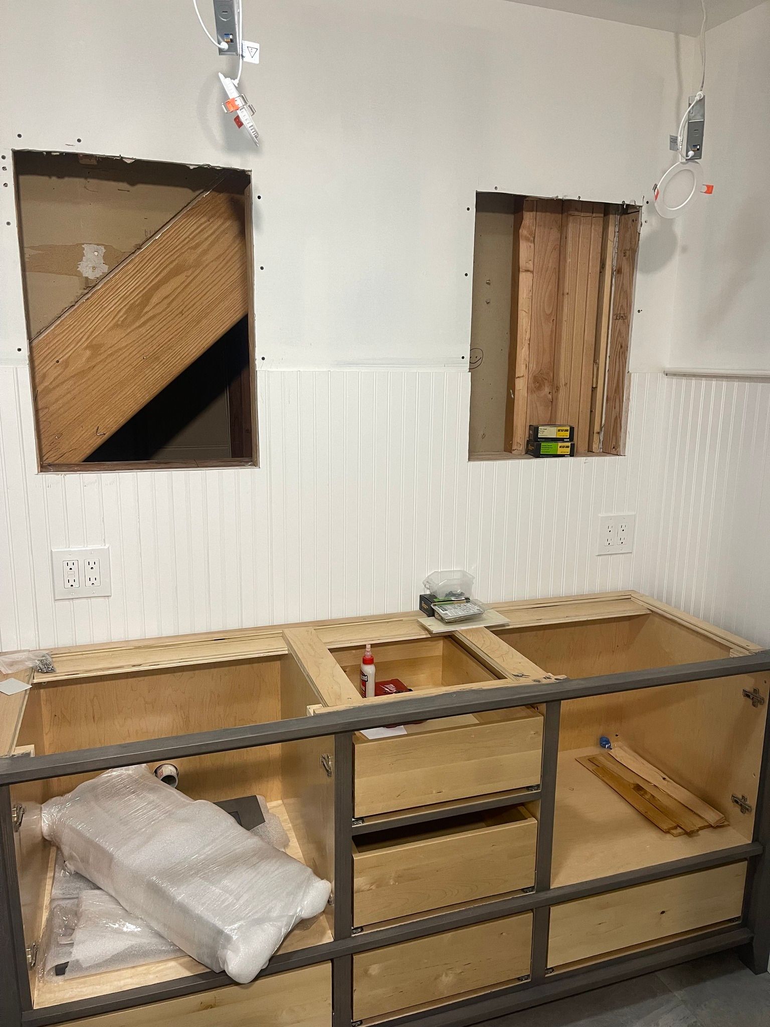 A bathroom renovation in progress with an unfinished vanity cabinet installed against a wall with two cut-out openings.