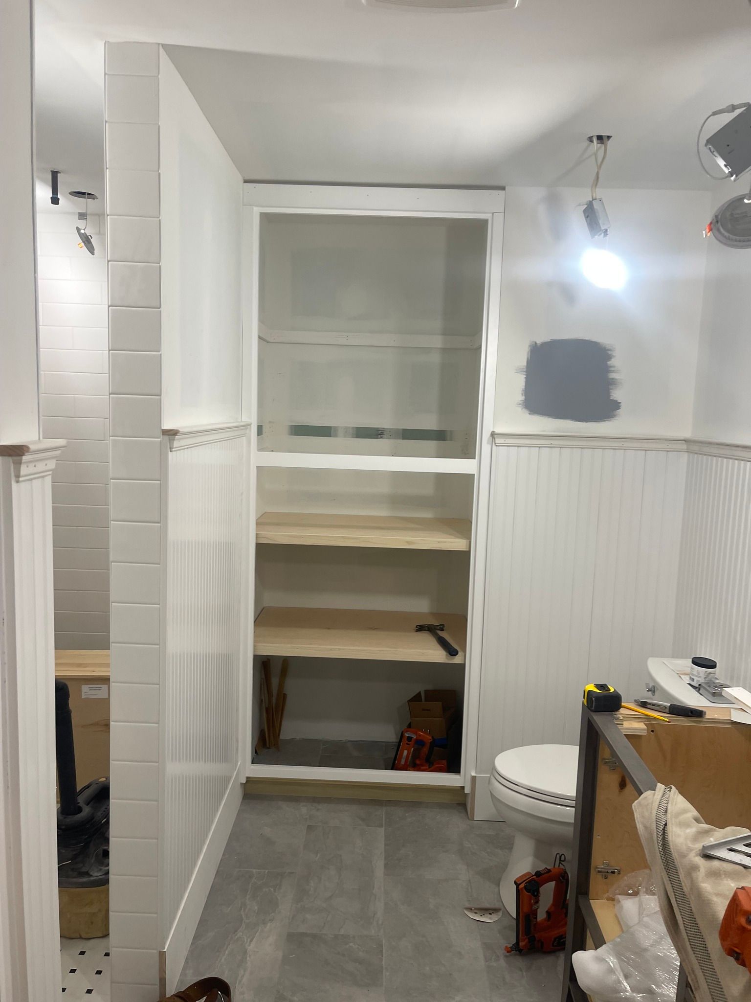 A bathroom under construction with white beadboard walls, a gray-tiled floor, open shelving, and a toilet.