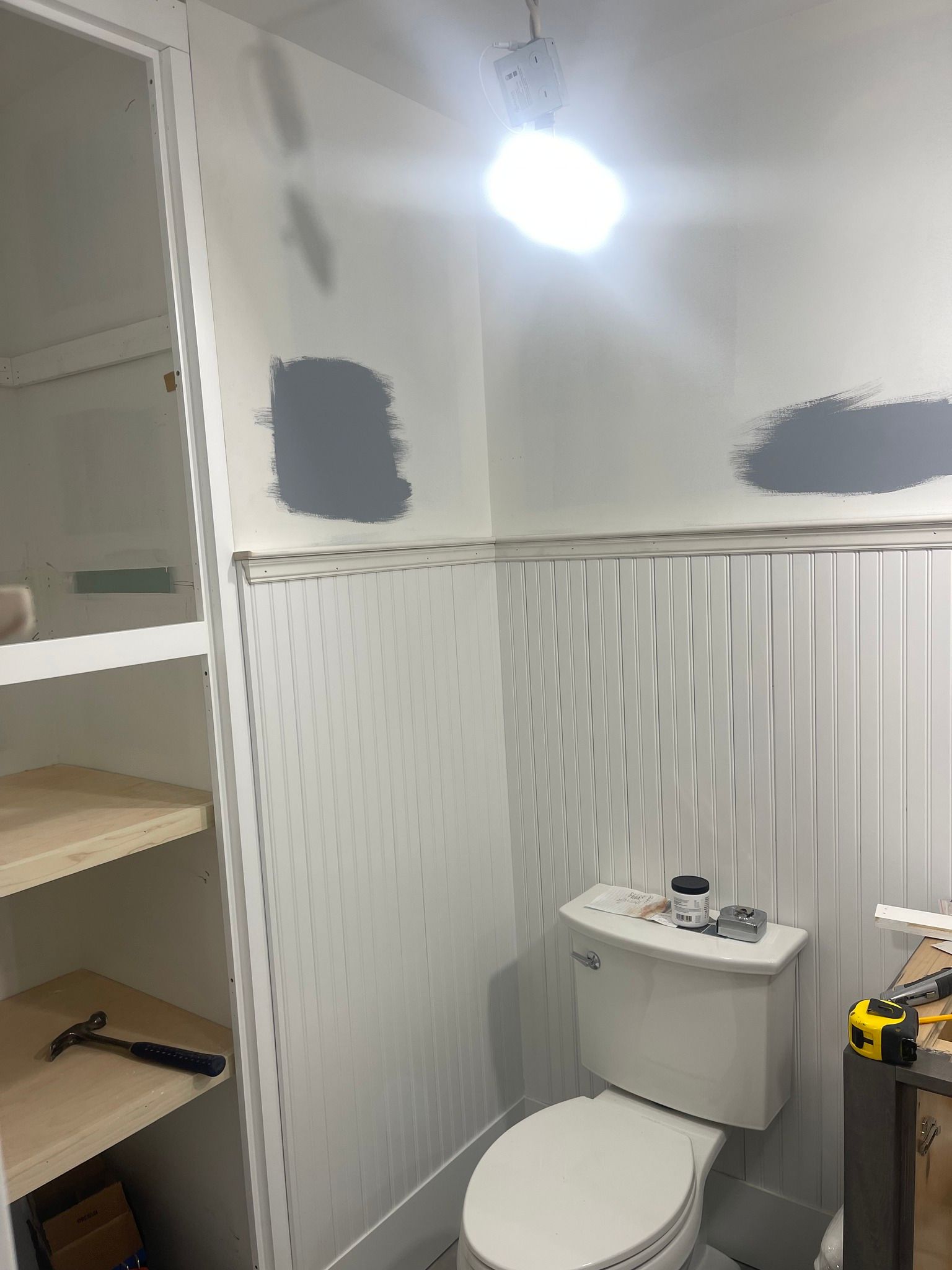 A bathroom undergoing renovation with white beadboard walls, grey paint samples, a toilet, and tools on shelves.