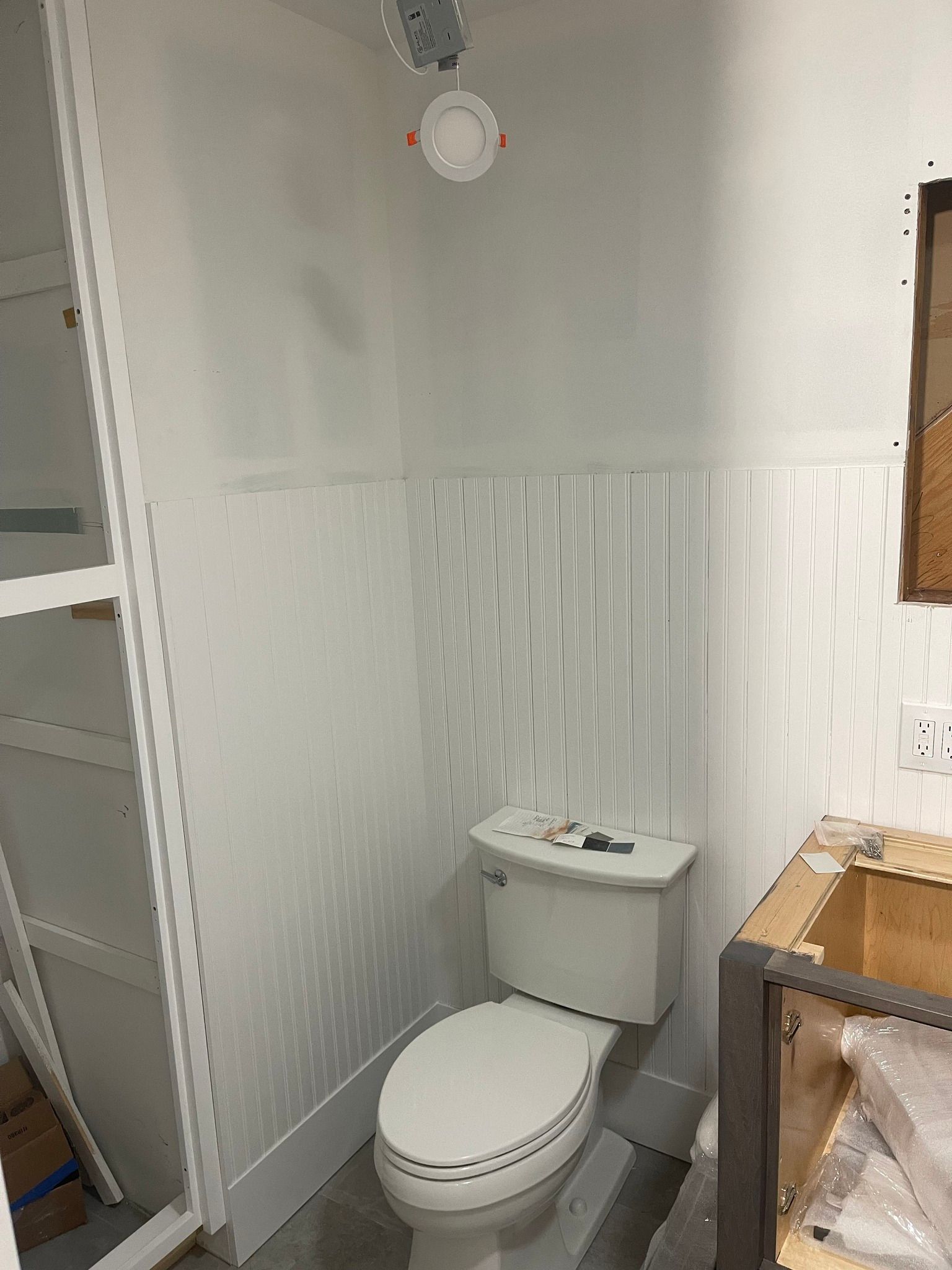An unfinished bathroom featuring white vertical wall paneling, a toilet, an exposed ceiling light, and a wooden vanity base.