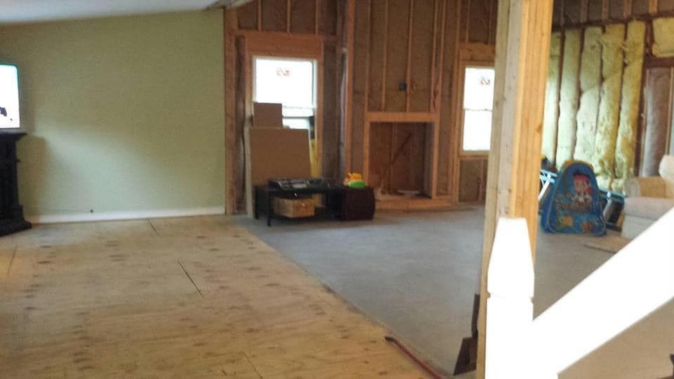 A room under renovation with exposed wall studs, insulation, a fireplace frame, and plywood flooring.