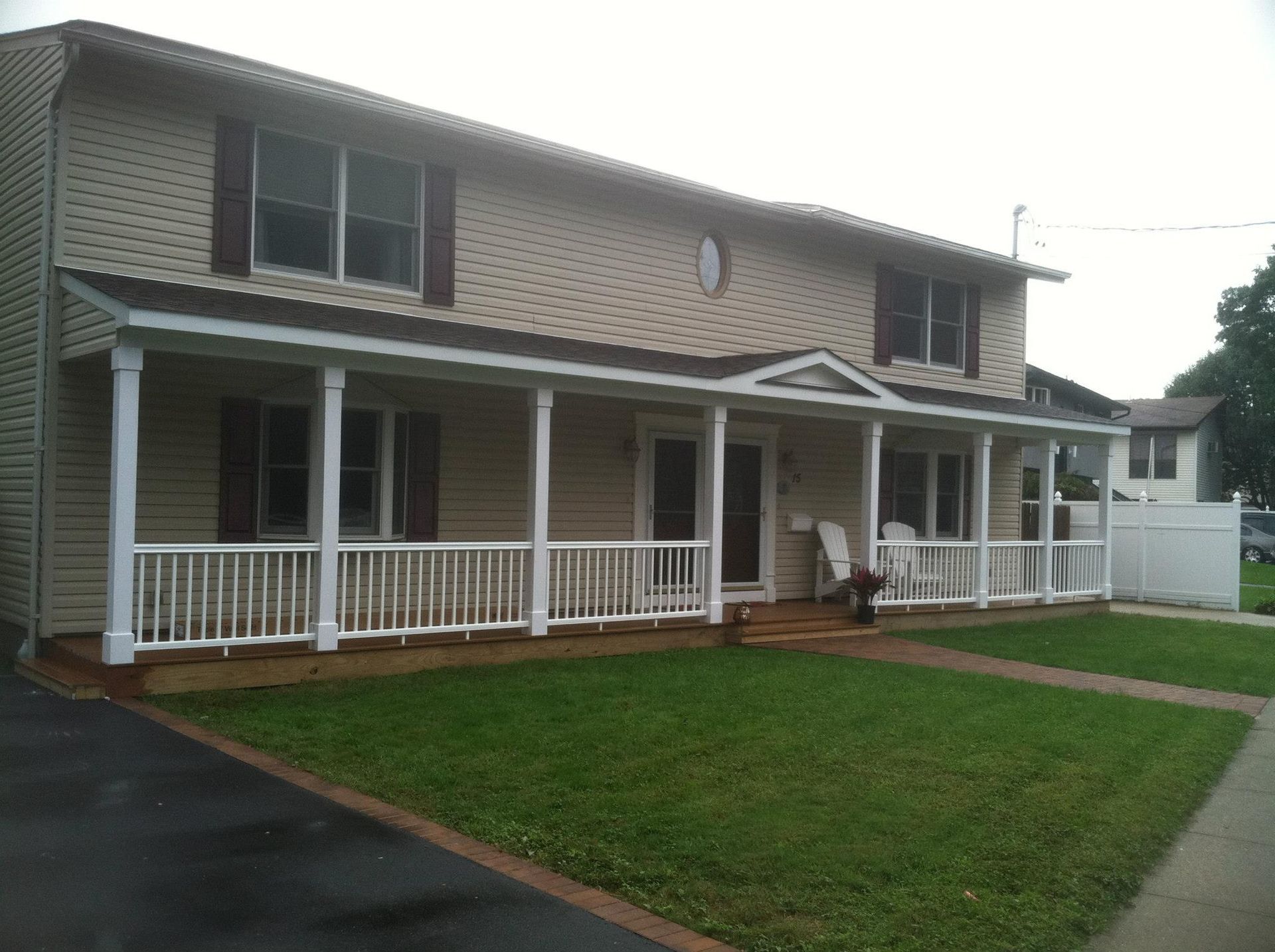 A two-story, beige suburban house with dark shutters, a large covered front porch, white railings, and a grassy front lawn.