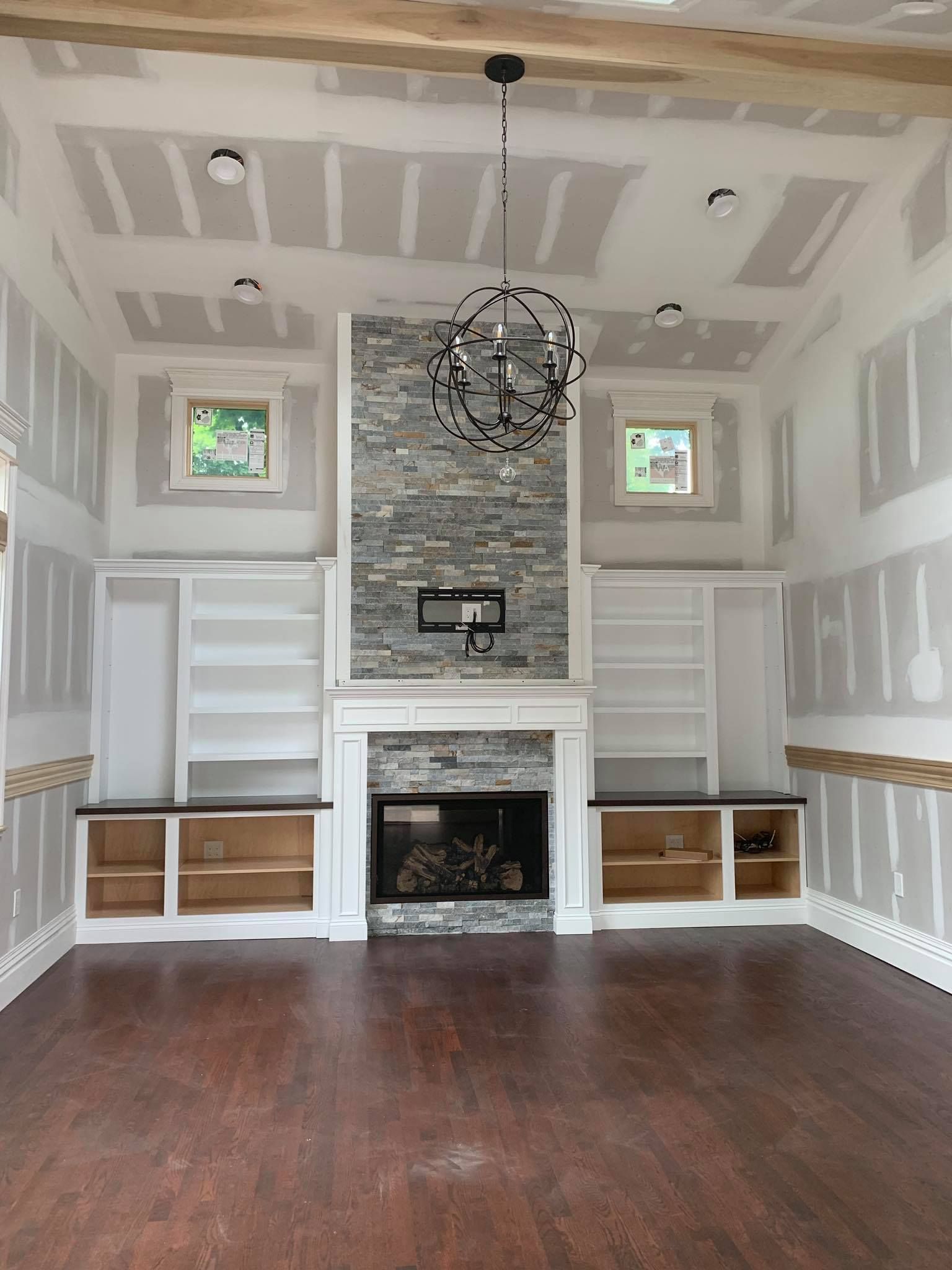 A room under construction with a stone fireplace, built-in shelving, hardwood floors, and unfinished drywall walls.