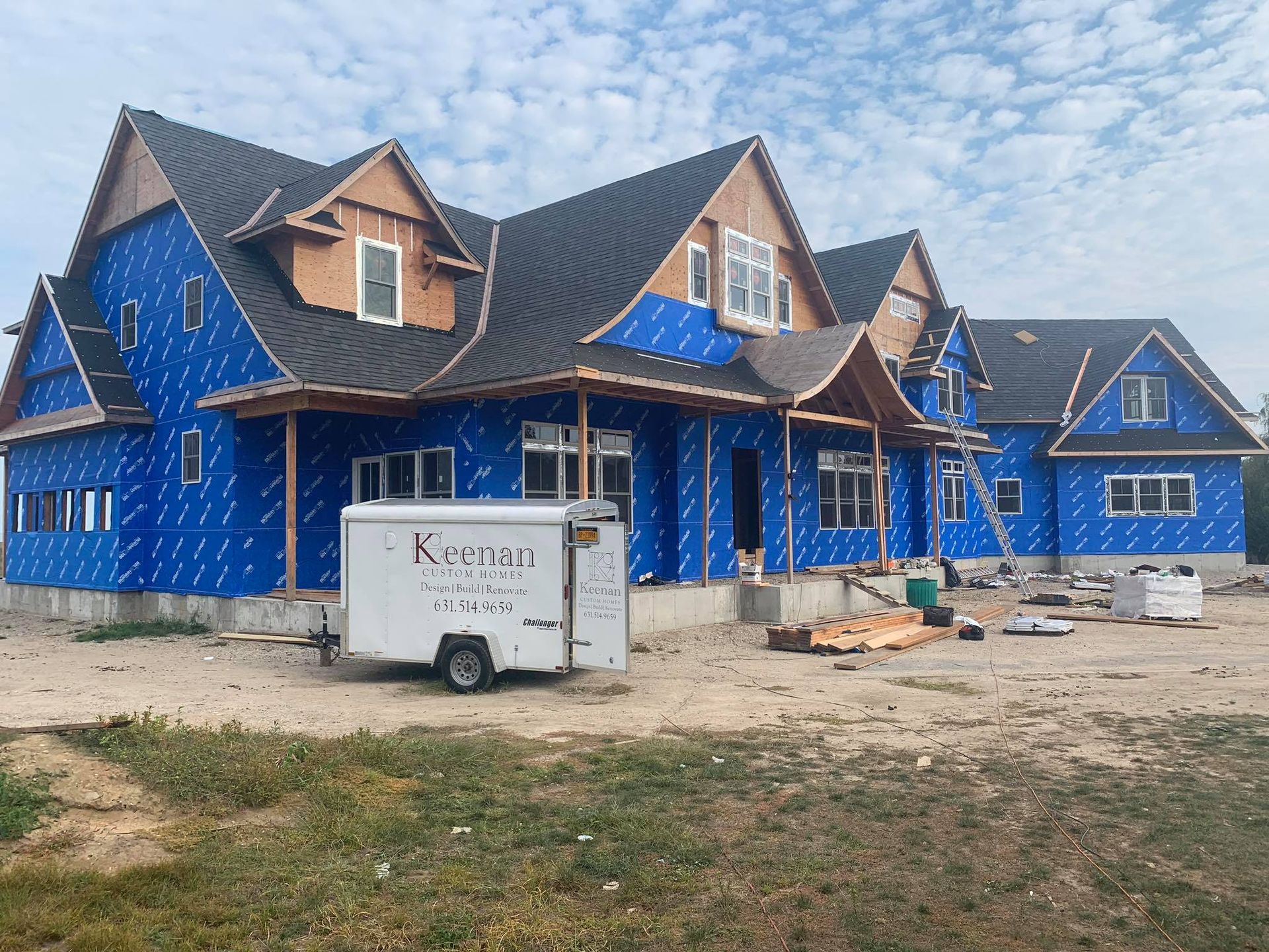 A large, multi-gabled house under construction, covered in blue protective wrap, with a white utility trailer in the yard.