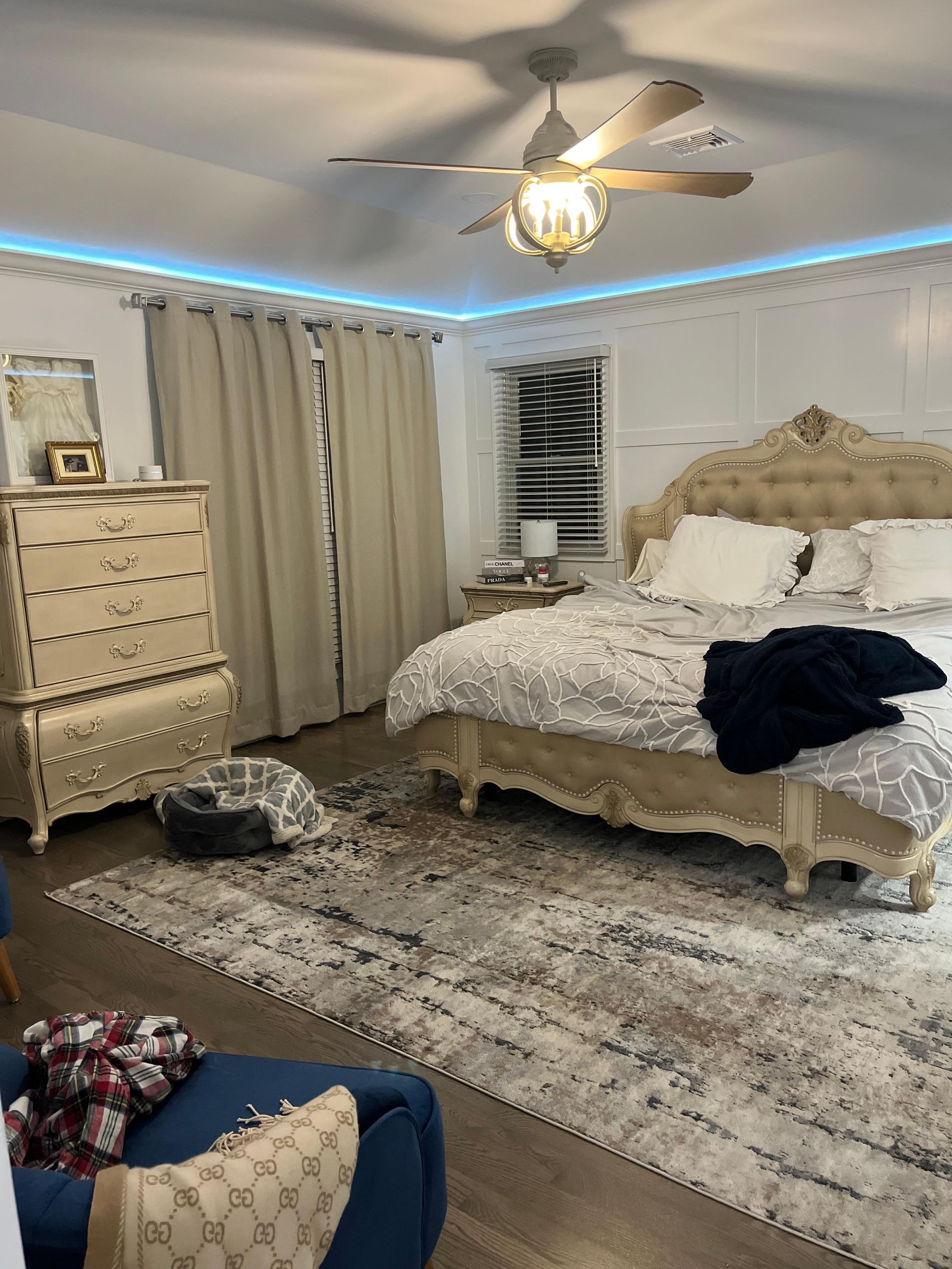 A bedroom with a tufted beige bed, patterned rug, tall dresser, tan curtains, and a glowing blue LED ceiling border.