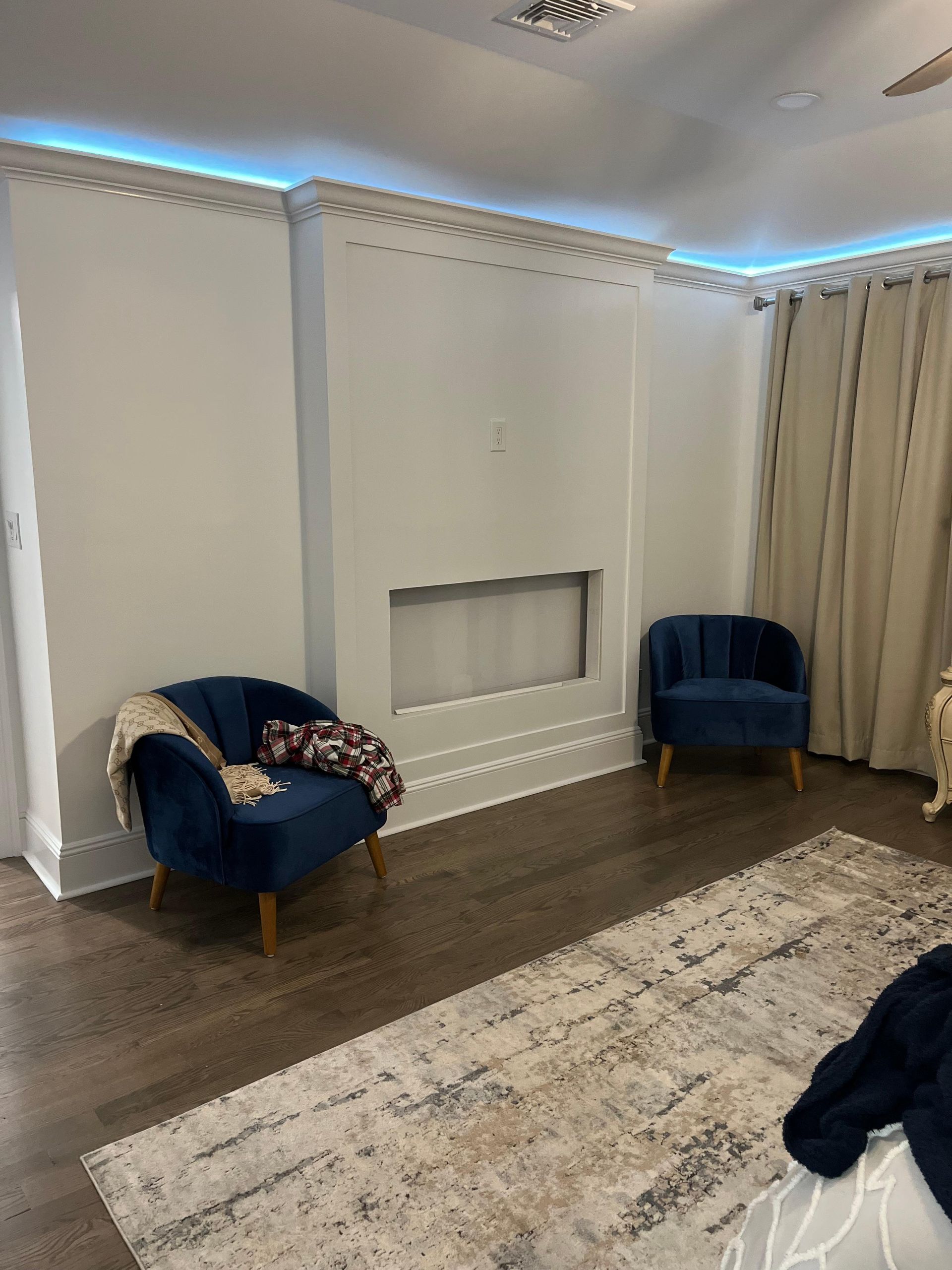 Two blue velvet chairs sit on a hardwood floor in front of a white wall with an inset rectangular panel and blue lighting.