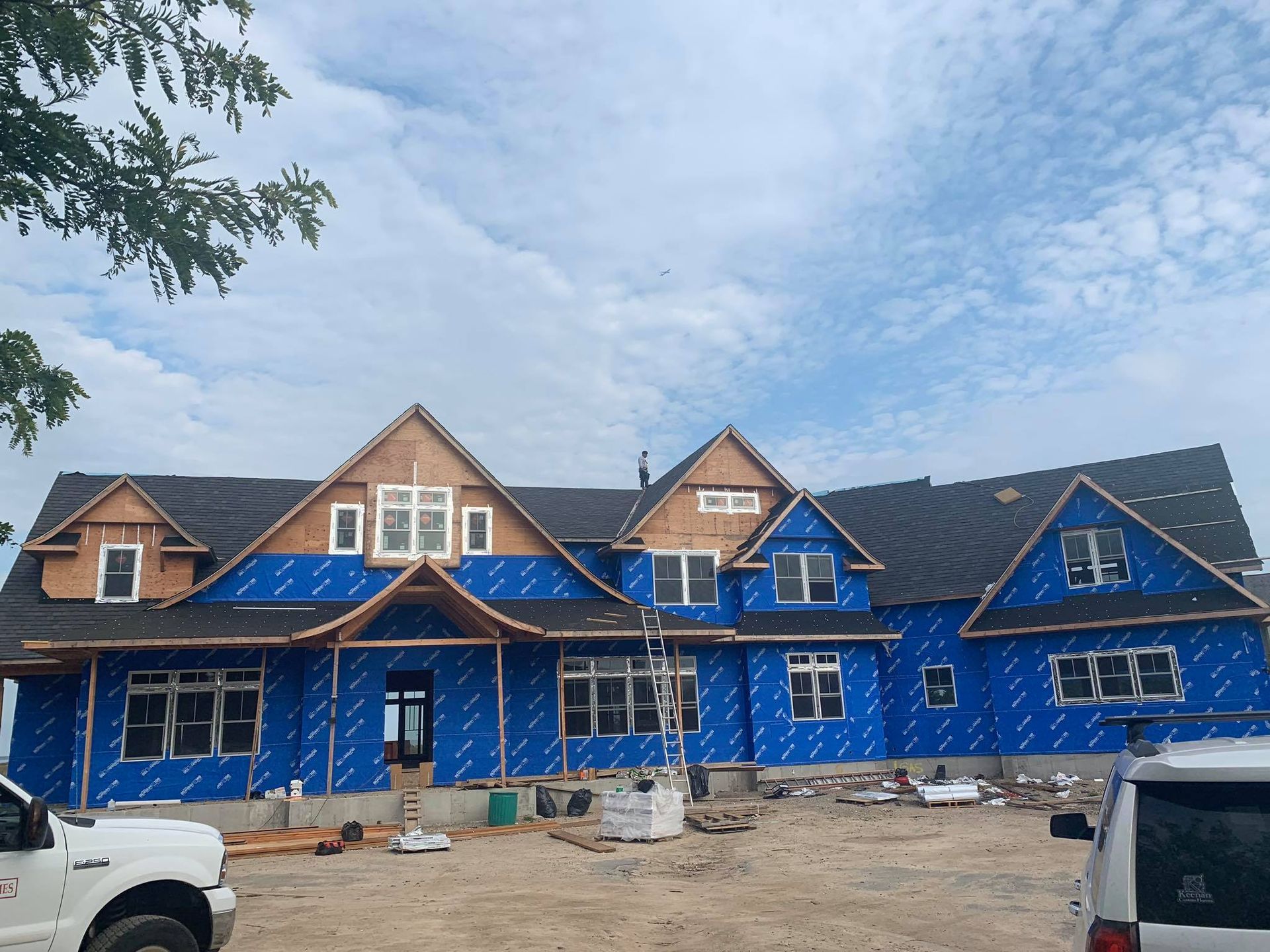 A large house under construction, covered in blue protective wrap, with shingled roofs and partially installed windows.