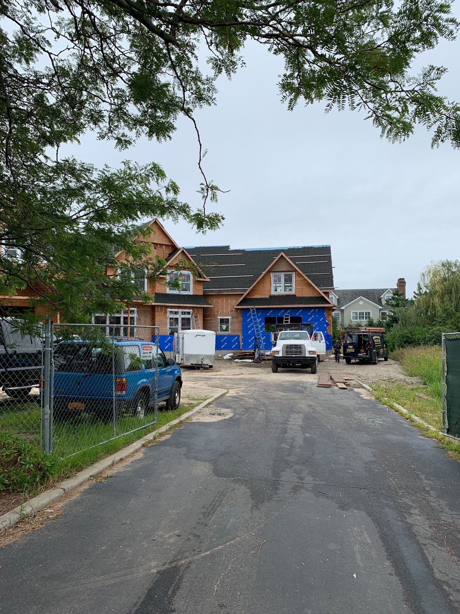 A house under construction with exposed wooden framing and blue sheathing, seen from a paved driveway.