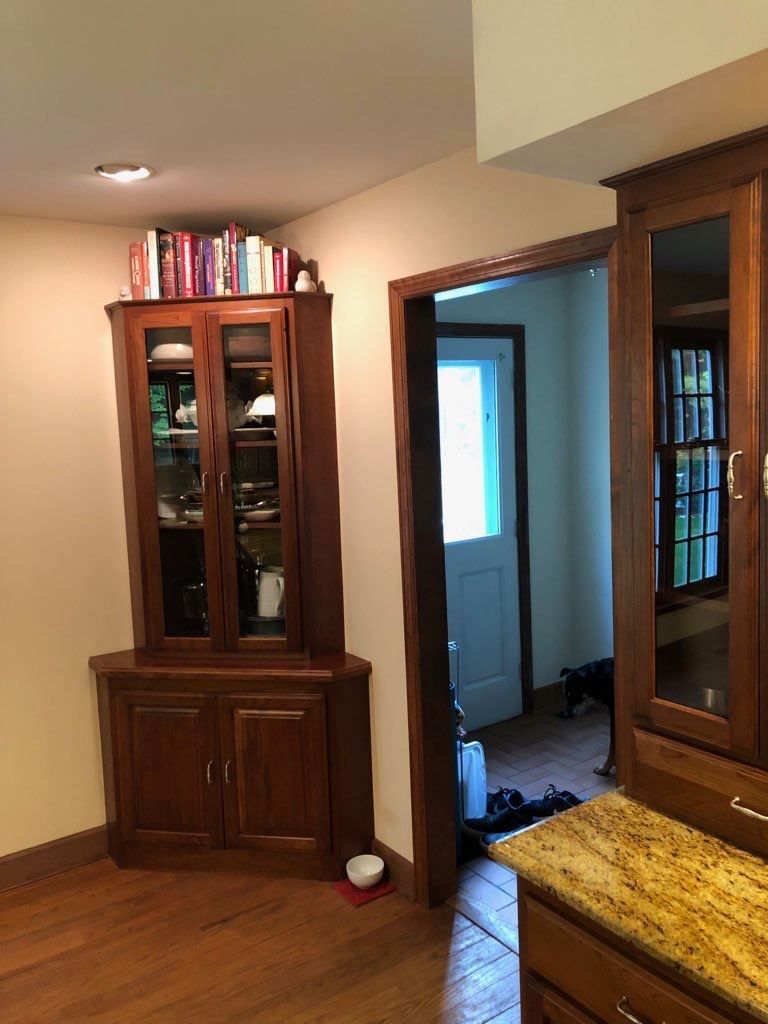 A wooden corner cabinet with glass doors stands in a room next to a doorway, with a small white bowl on the floor.