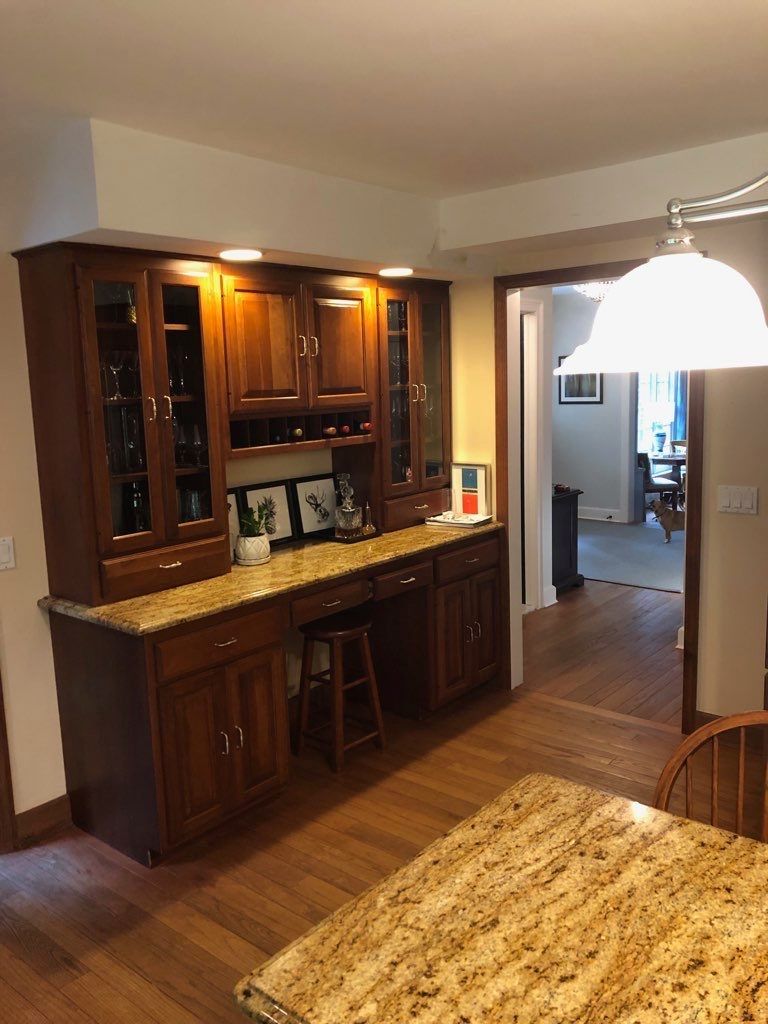 A wooden hutch with glass doors and drawers, featuring a granite countertop and a stool in a dining area.