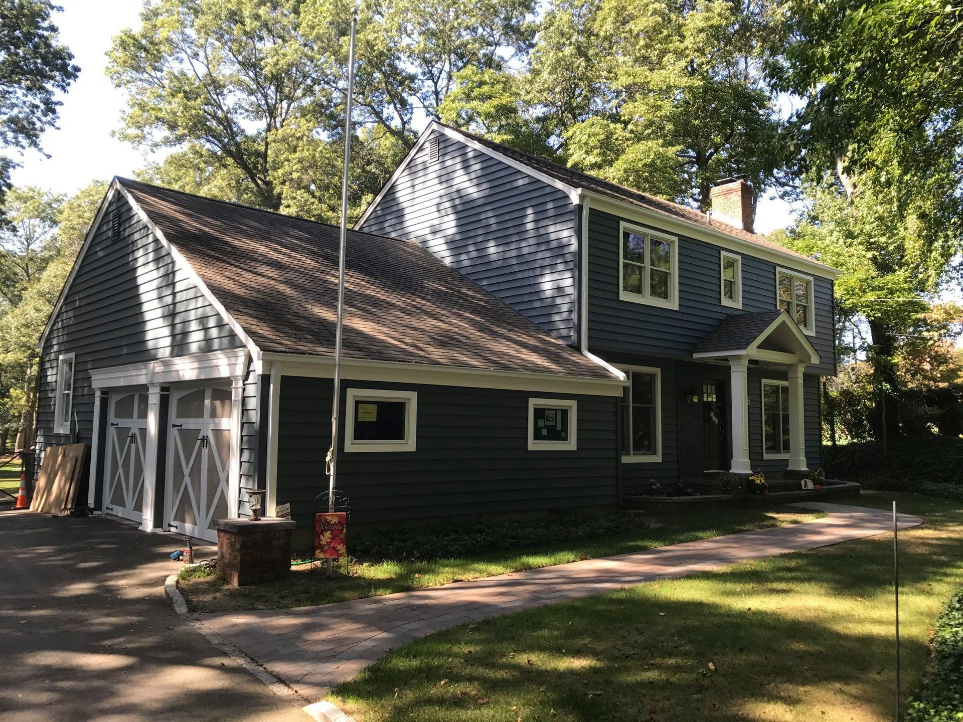 A two-story dark blue house with white trim, a garage, and a front walkway, set among trees on a sunny day.