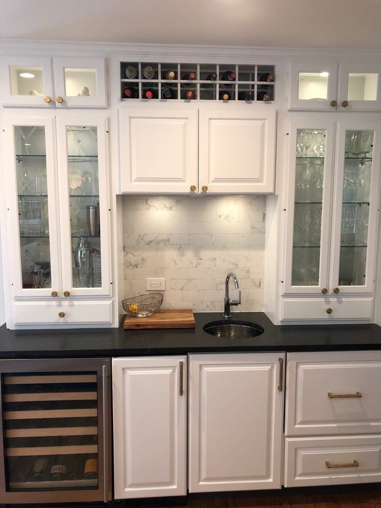 A white wet bar with glass-front cabinets, a wine rack, black countertops, a sink, and a built-in wine cooler.