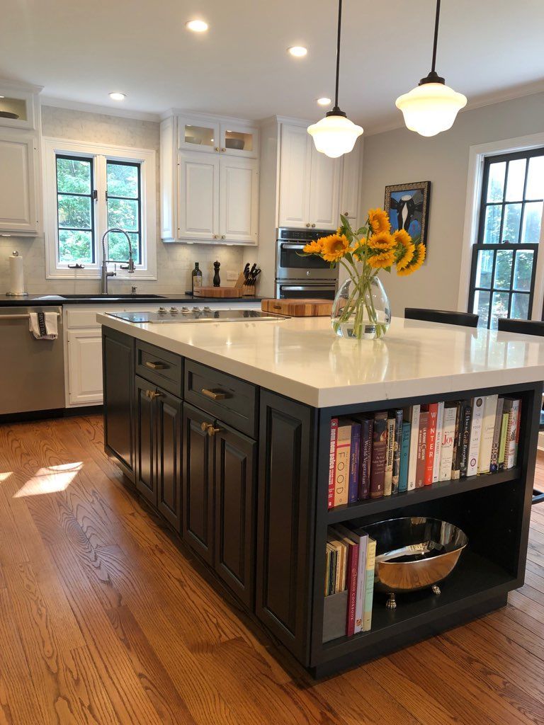 A modern kitchen featuring a dark blue island with built-in bookshelves and sunflowers, white cabinets, and wood floors.
