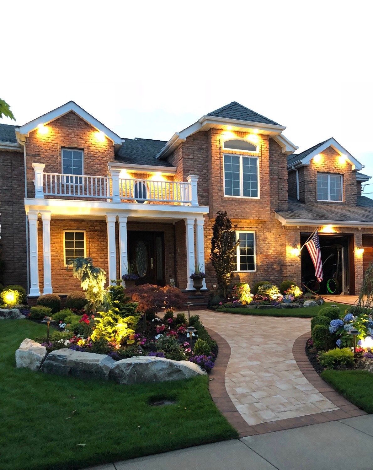 A large, multi-story brick house at dusk, with illuminated windows, a paved walkway, and landscaping in the front yard.