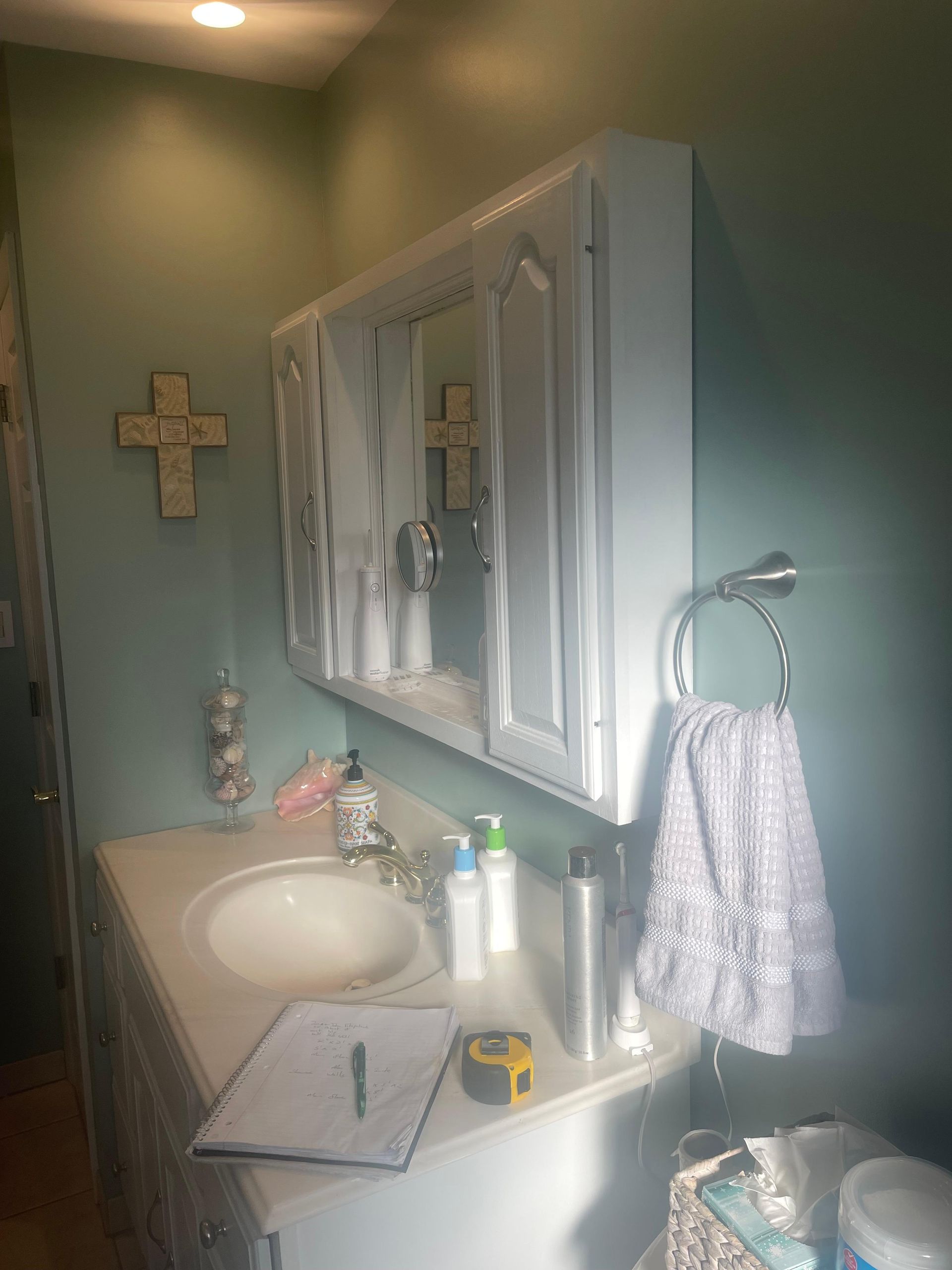 A white bathroom cabinet with a mirror above a sink, with a hand towel on a ring and a notepad on the counter.