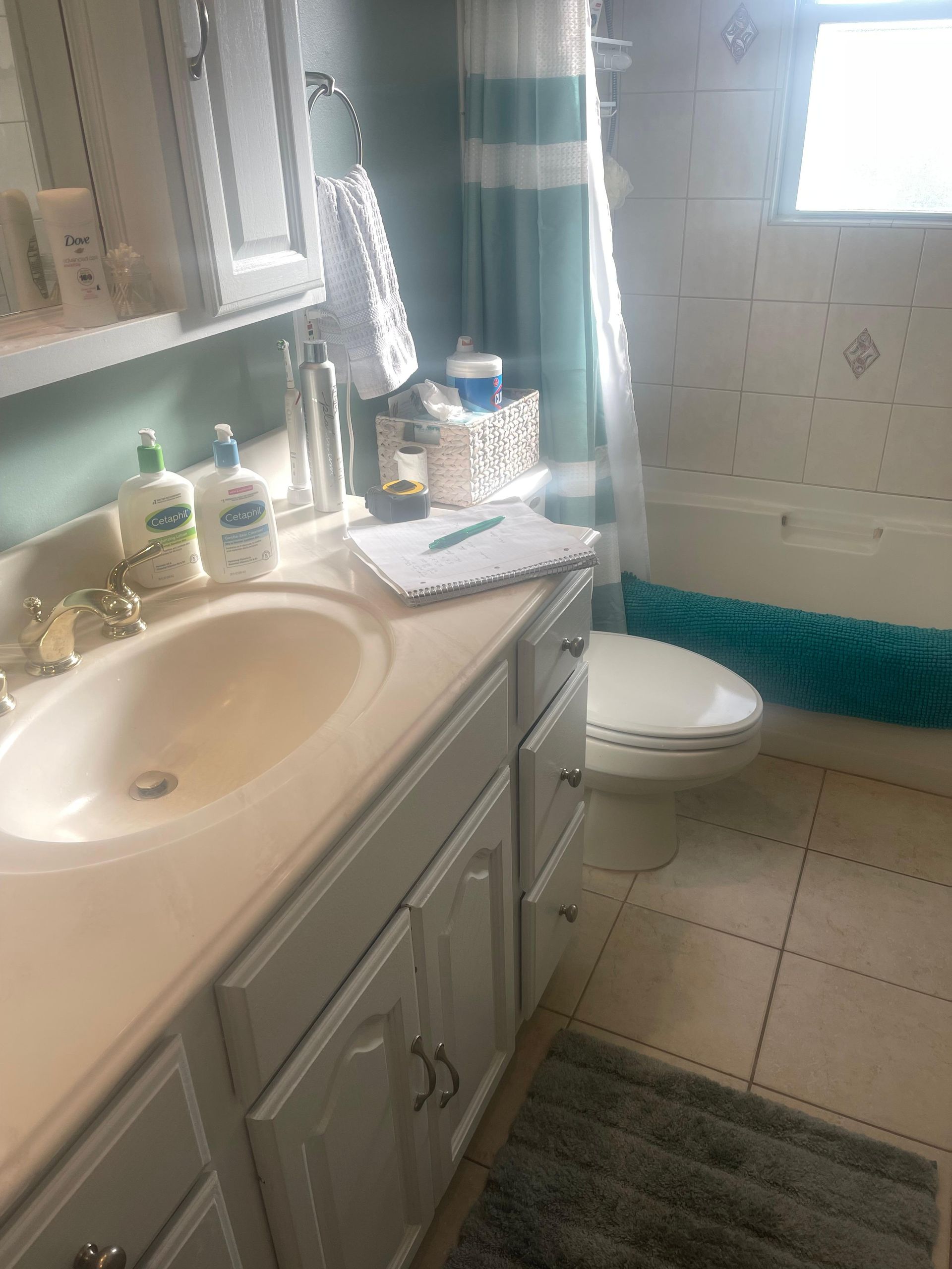 A bright bathroom with a light-colored vanity sink, a toilet, a teal shower curtain, and beige tile flooring.