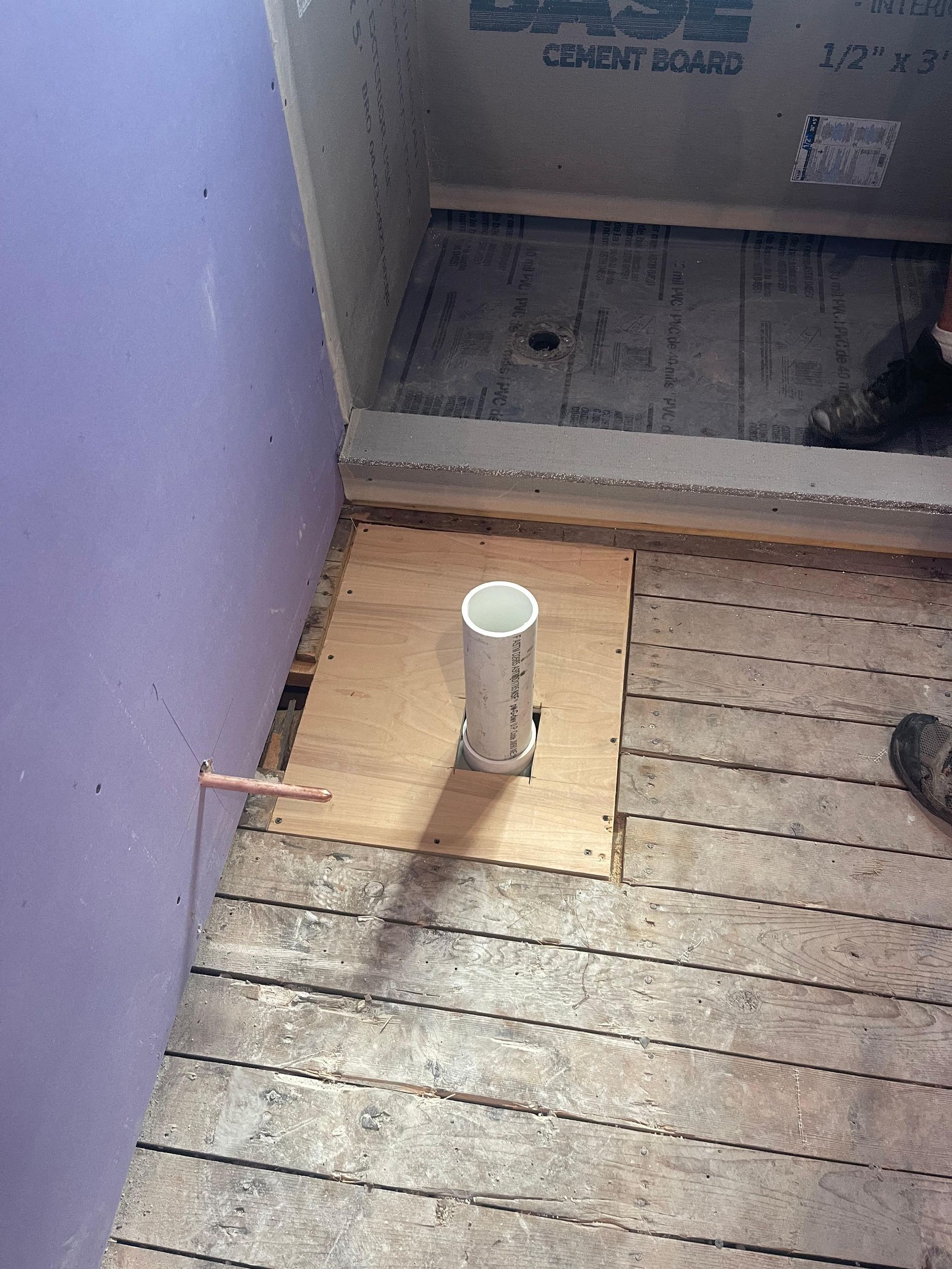A white PVC drain pipe sticks up from a plywood patch in a subfloor next to a purple drywall wall and a shower area.