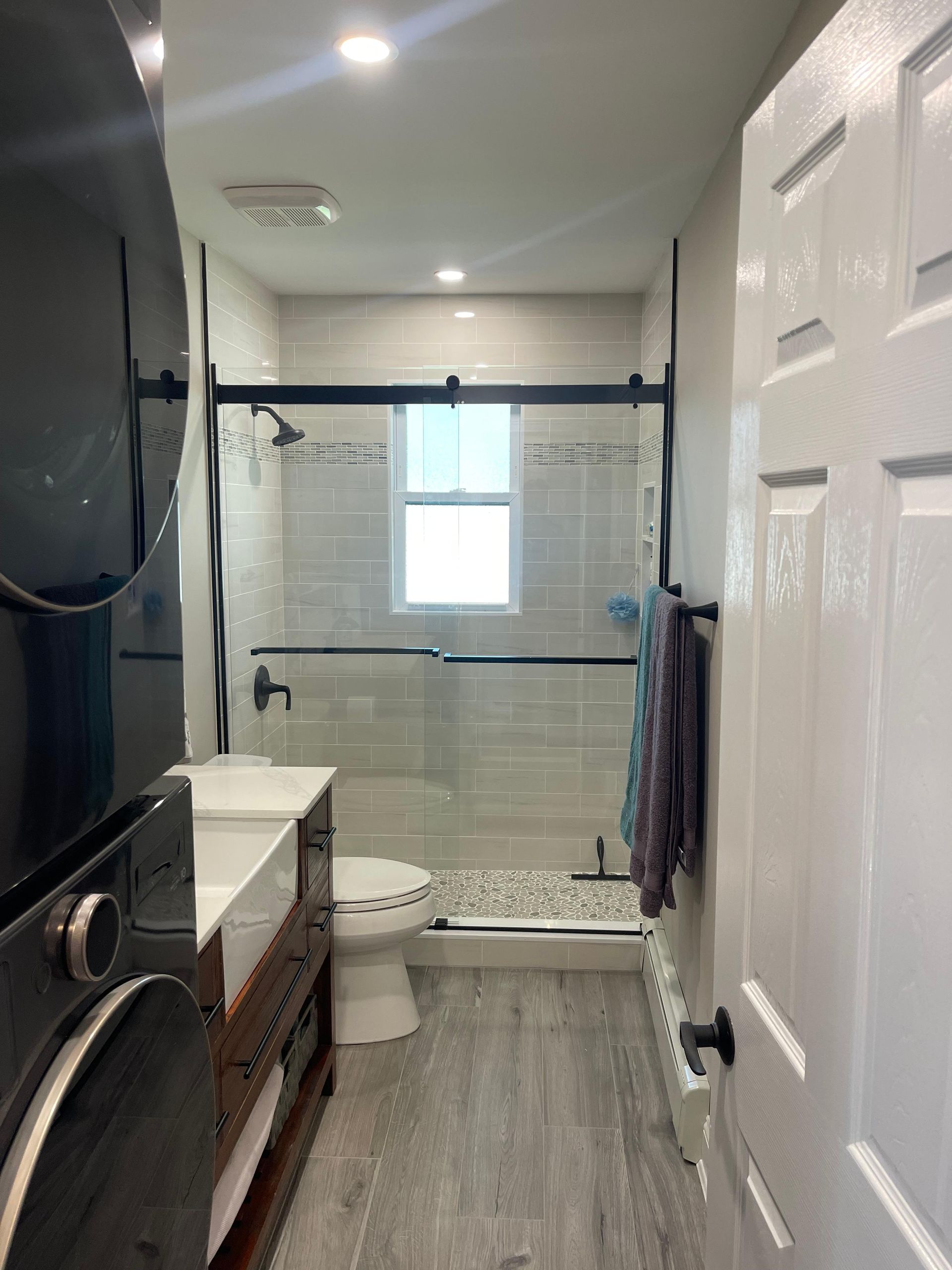 A narrow bathroom featuring a stacked washer-dryer, a wood vanity, a toilet, and a glass-enclosed shower with tiled walls.