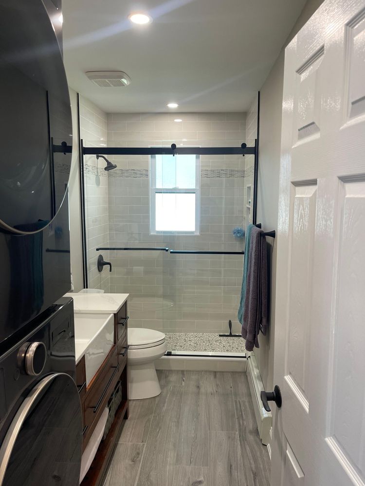 A narrow bathroom featuring a stacked washer-dryer unit, a wooden vanity, a white toilet, and a glass-enclosed shower.