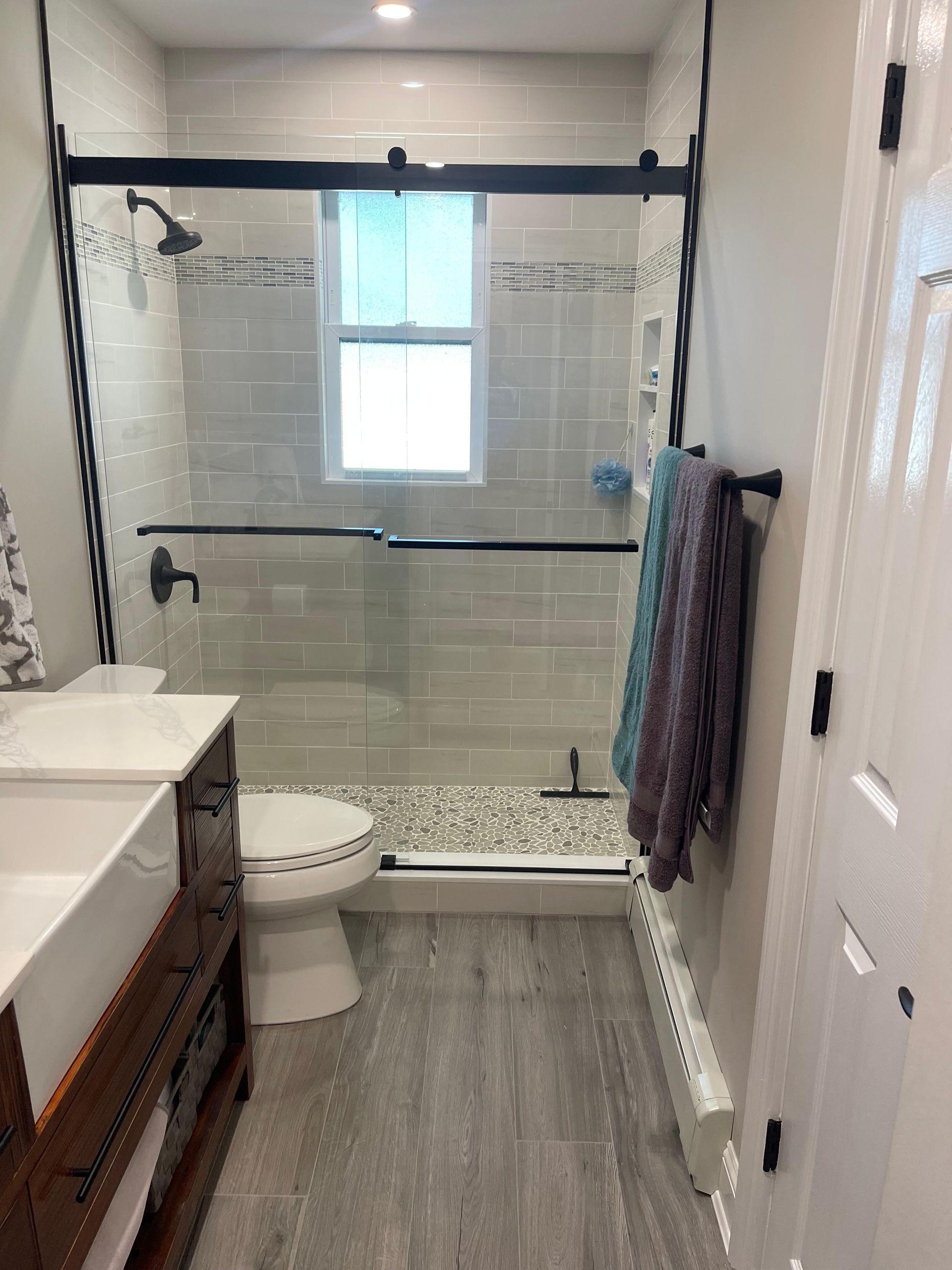A modern bathroom featuring a white vanity, toilet, and glass-enclosed shower with subway tiles and grey wood-look flooring.