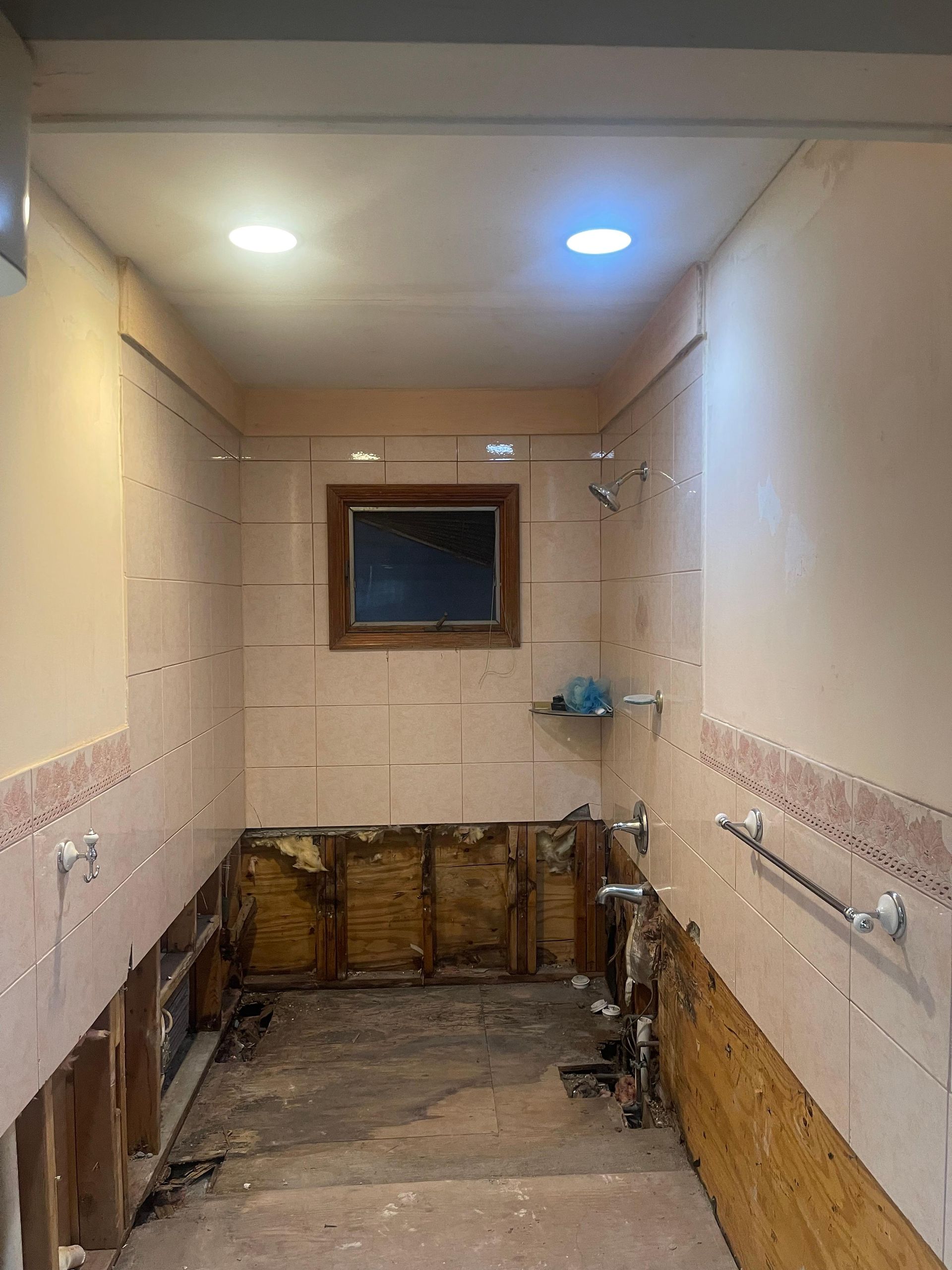 A bathroom under renovation with exposed wall studs, removed vanity cabinets, and damaged drywall below the tile line.