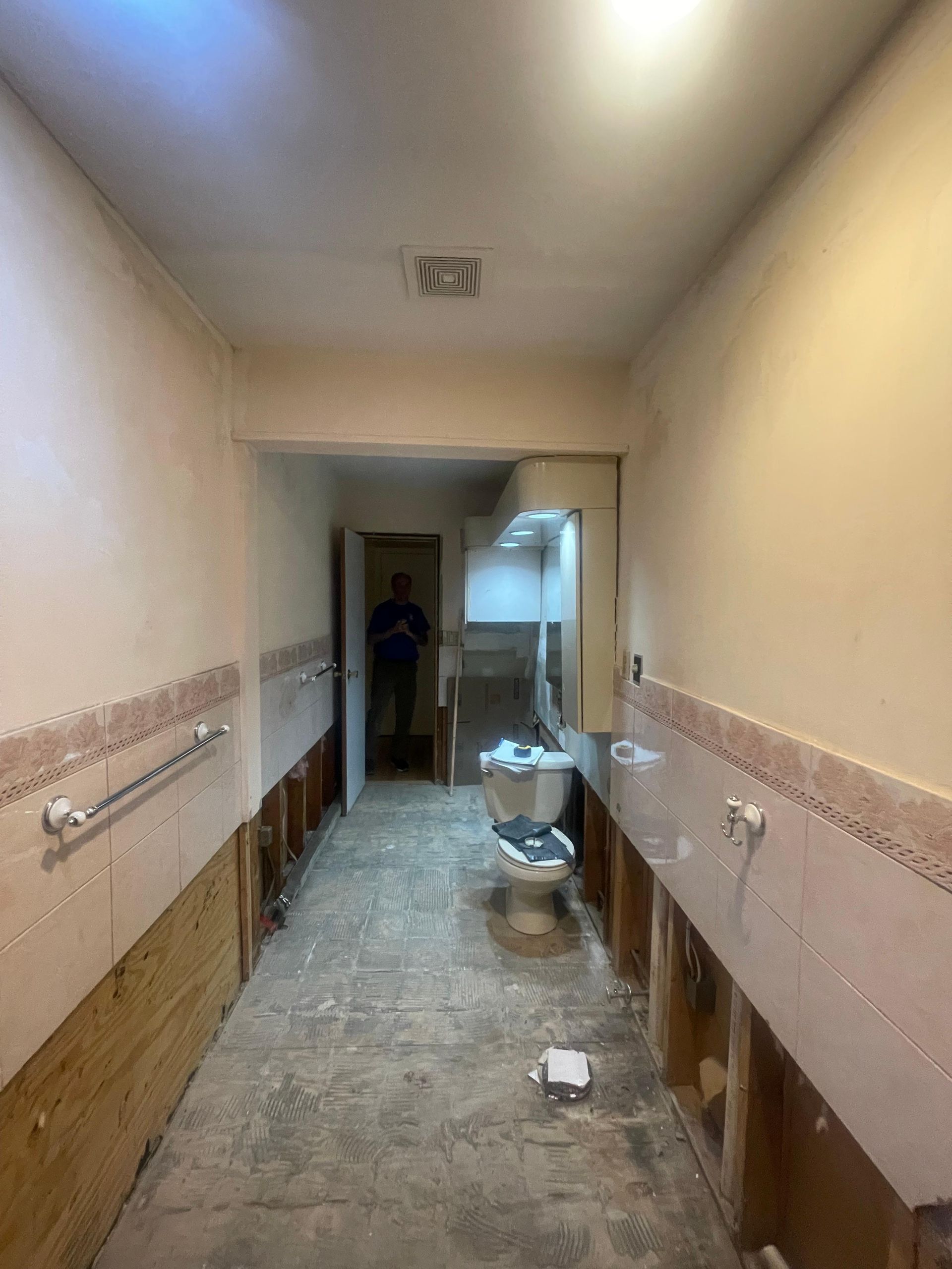 A hallway under renovation showing exposed wall studs, removed baseboards, a single toilet, and a person in the distance.