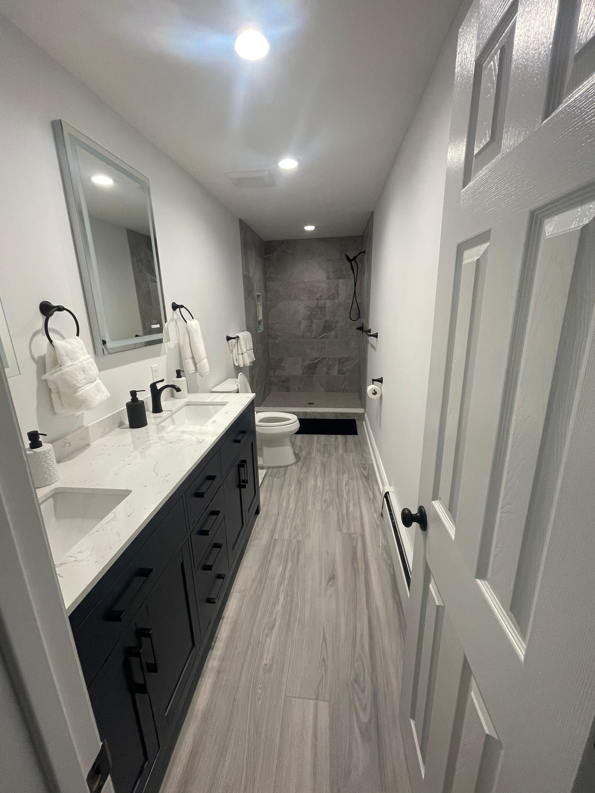 A long, modern bathroom with a dark vanity, white countertop, gray wood-look flooring, and a walk-in shower at the end.
