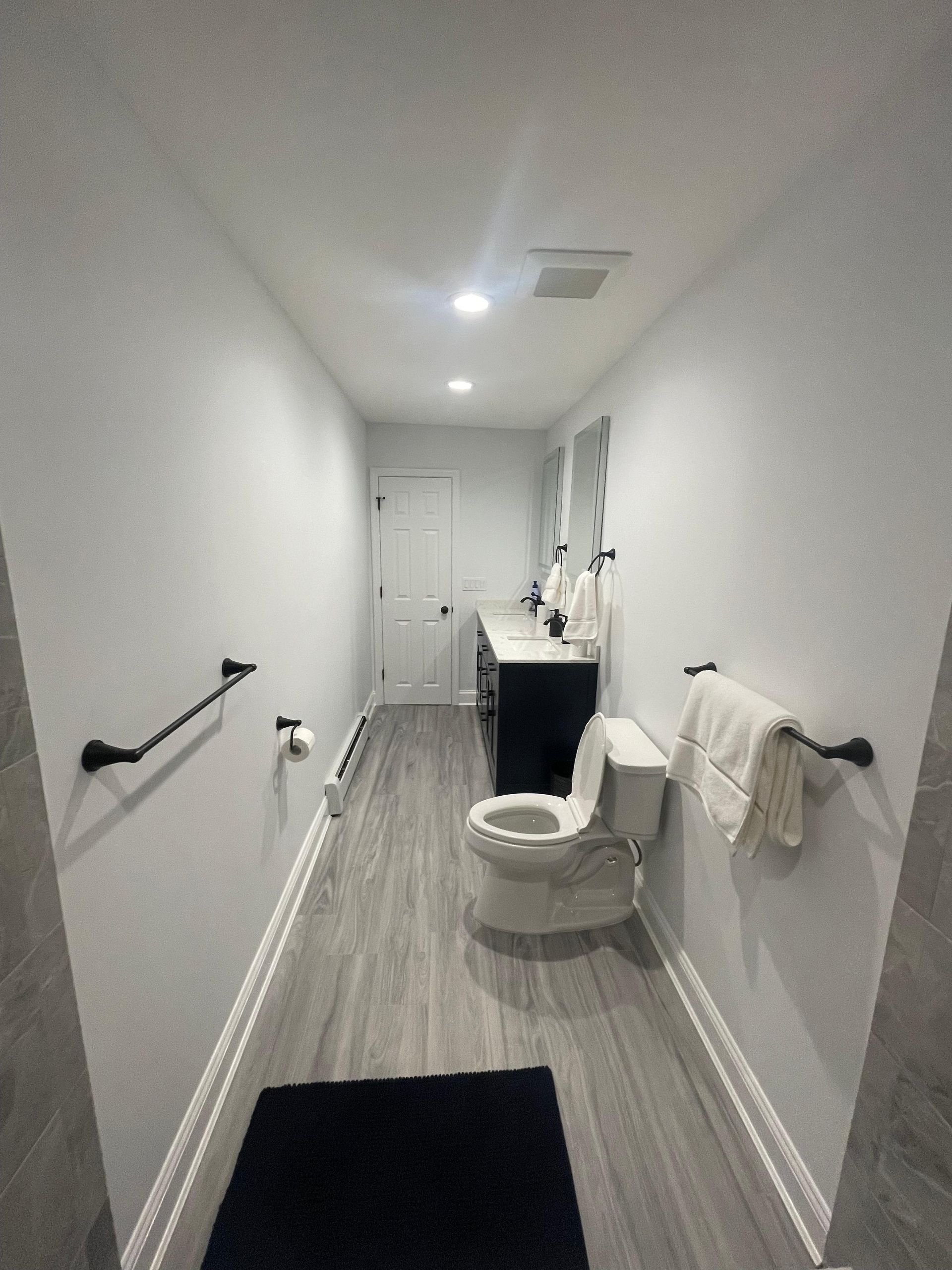 A narrow, modern bathroom with light gray walls, wood-look flooring, a white toilet, and dark vanity with dual mirrors.