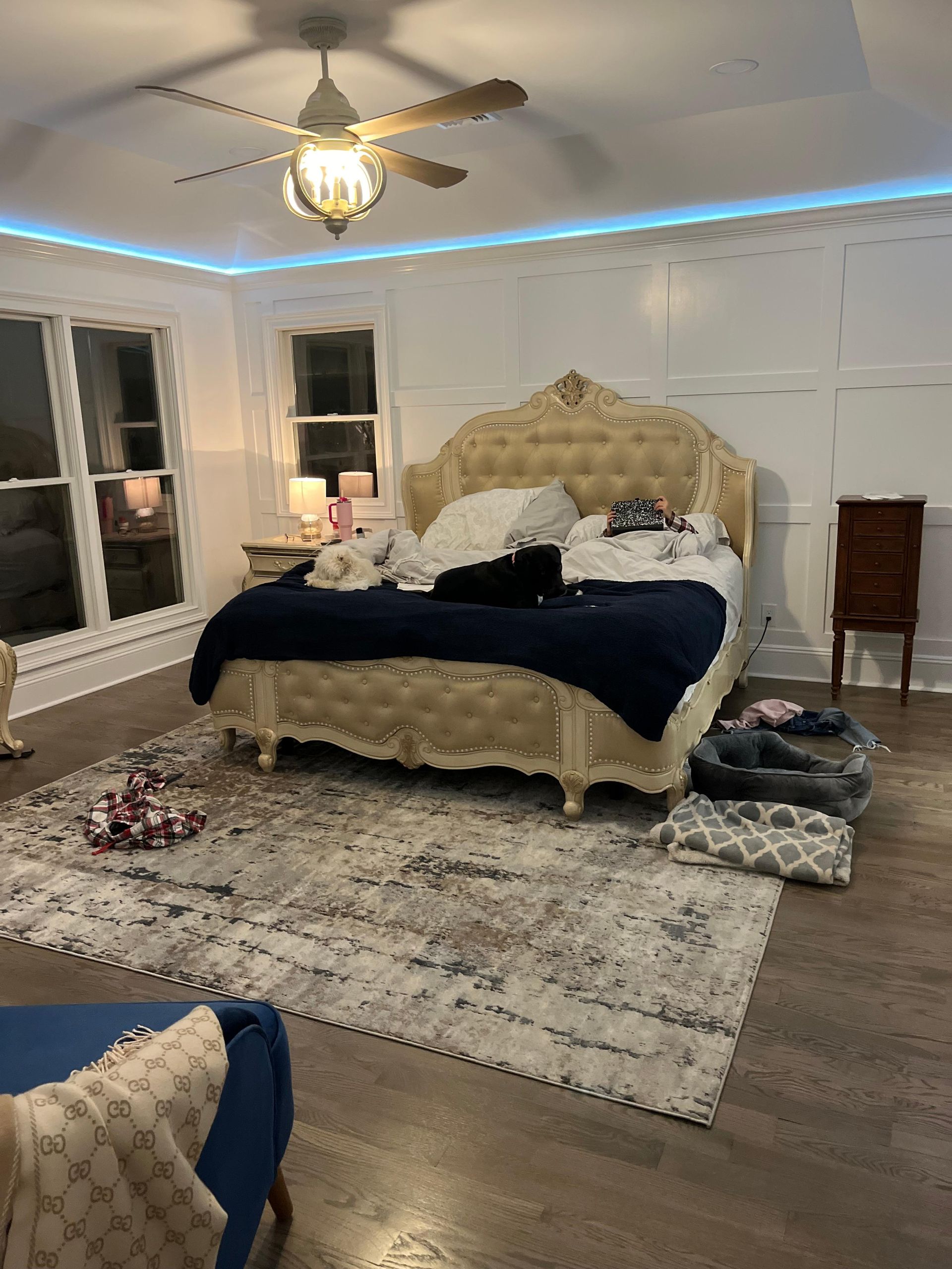 A large tufted beige bed with navy bedding in a bedroom with white paneled walls, a ceiling fan, and a textured rug.