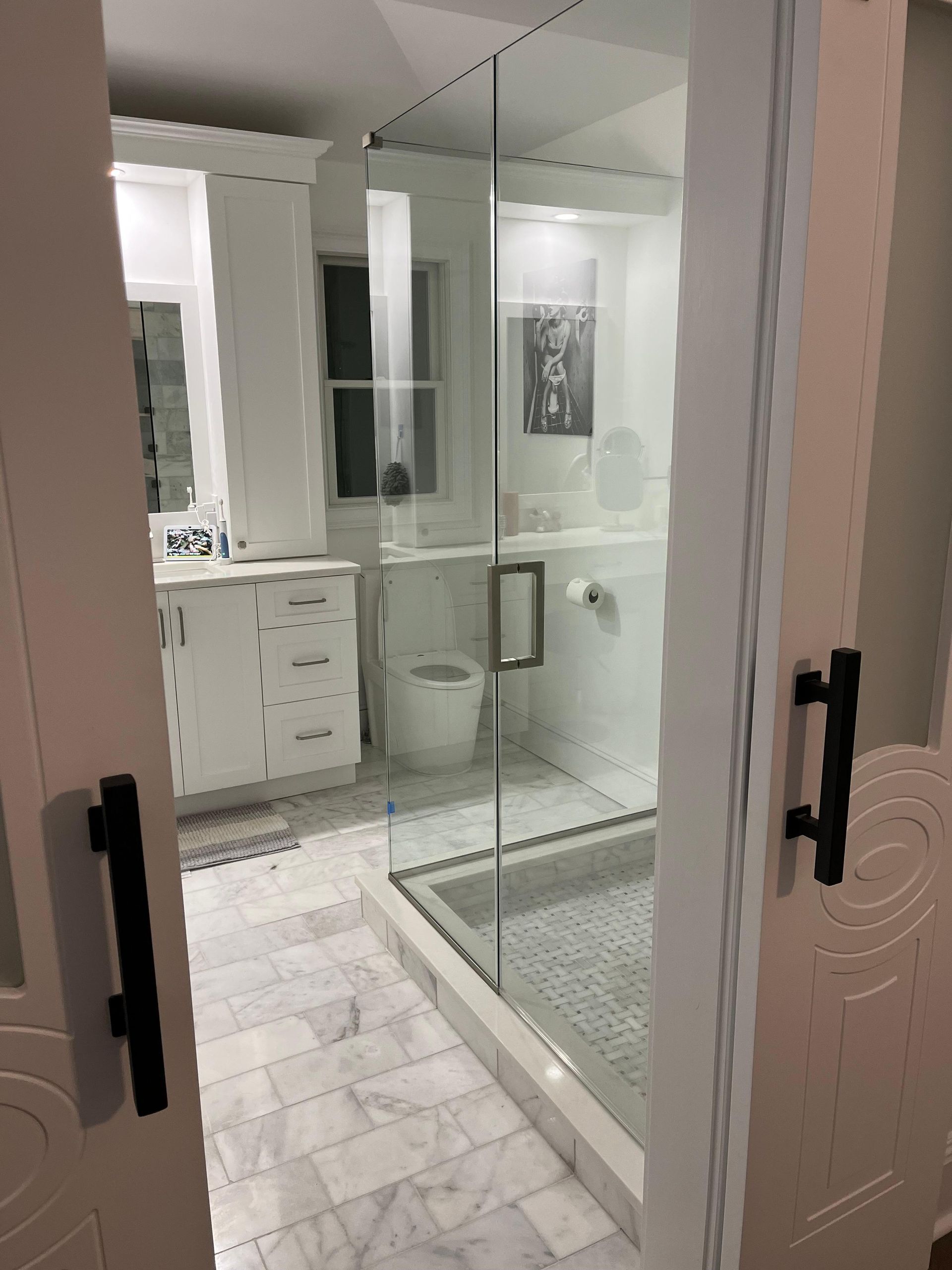 A bathroom viewed through white sliding doors, featuring a white vanity, marble tile floor, and a glass-enclosed shower.