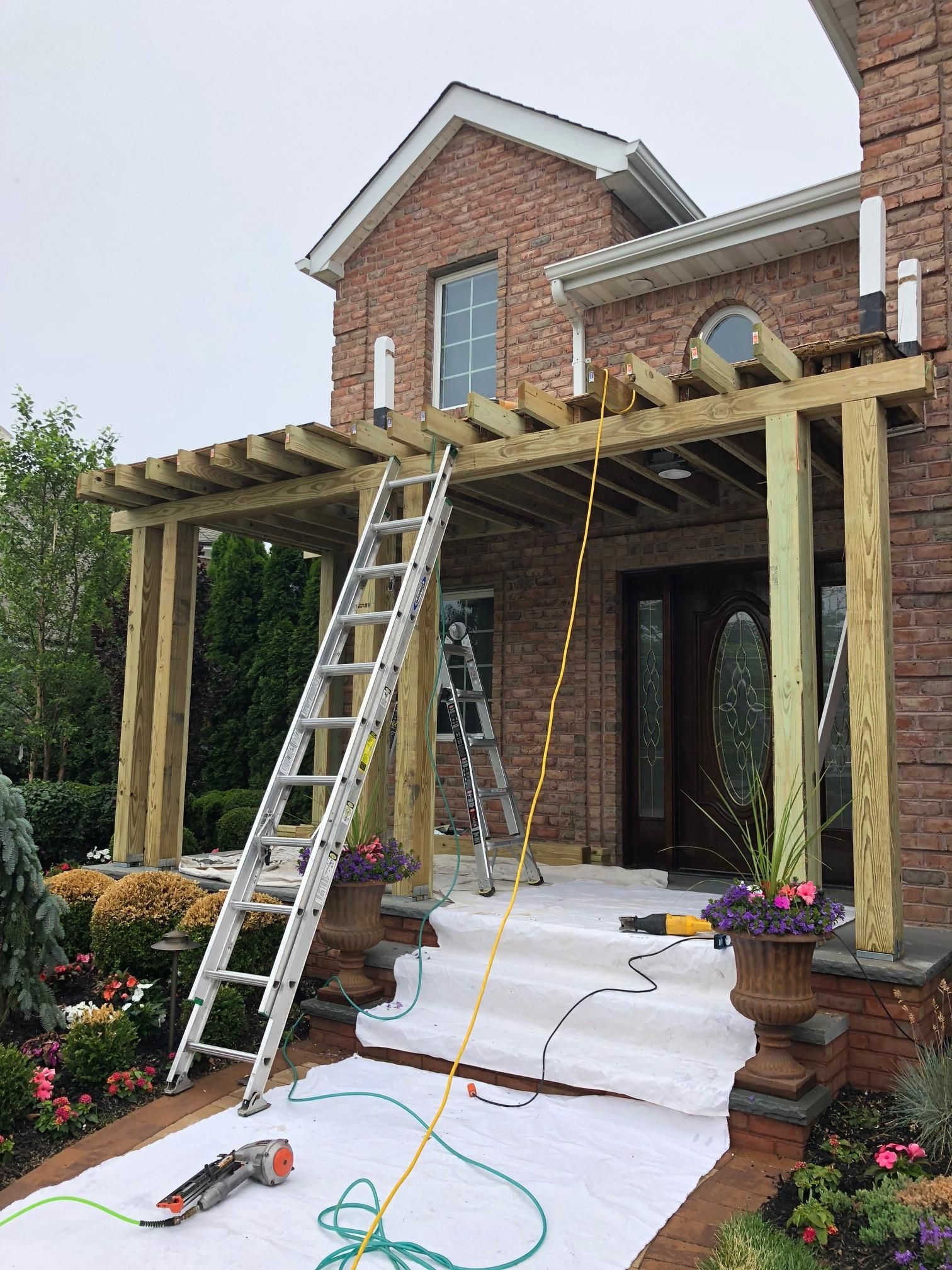 A wooden pergola under construction over a brick house entrance, with ladders and tools on a protective floor covering.