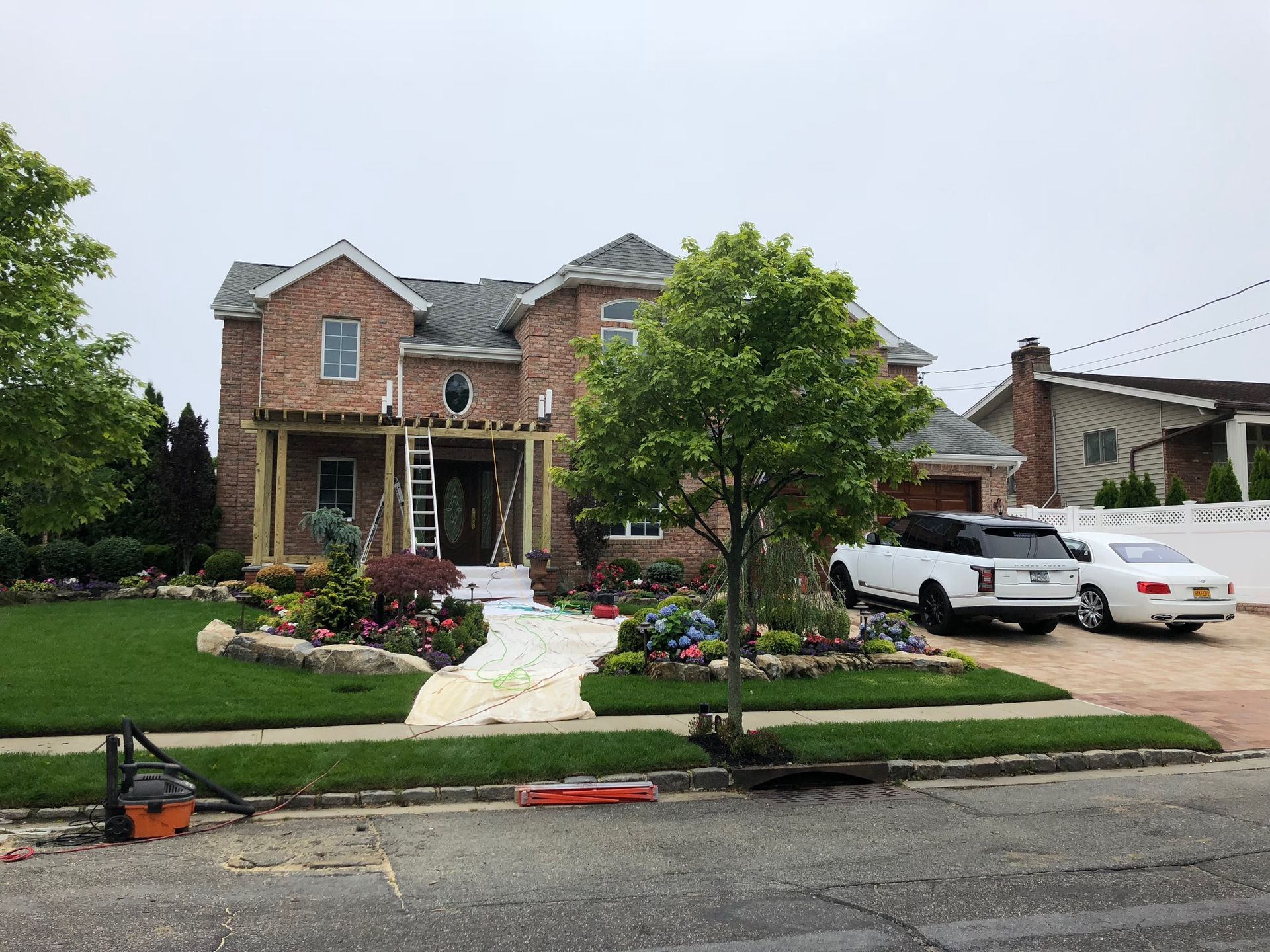 A brick two-story house with a partially constructed wooden porch frame, surrounded by landscaping, trees, and a driveway.