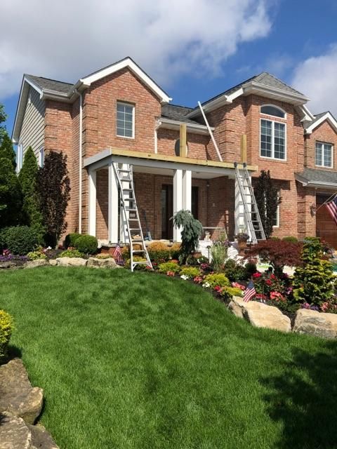 A brick house with a front porch under construction, featuring ladders and scaffolding, surrounded by landscaped gardens.