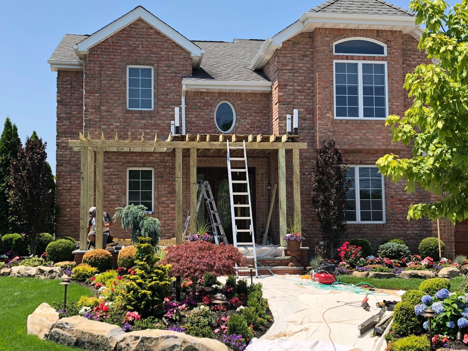 A construction worker builds a wooden pergola over the front entrance of a two-story brick house with a landscaped yard.