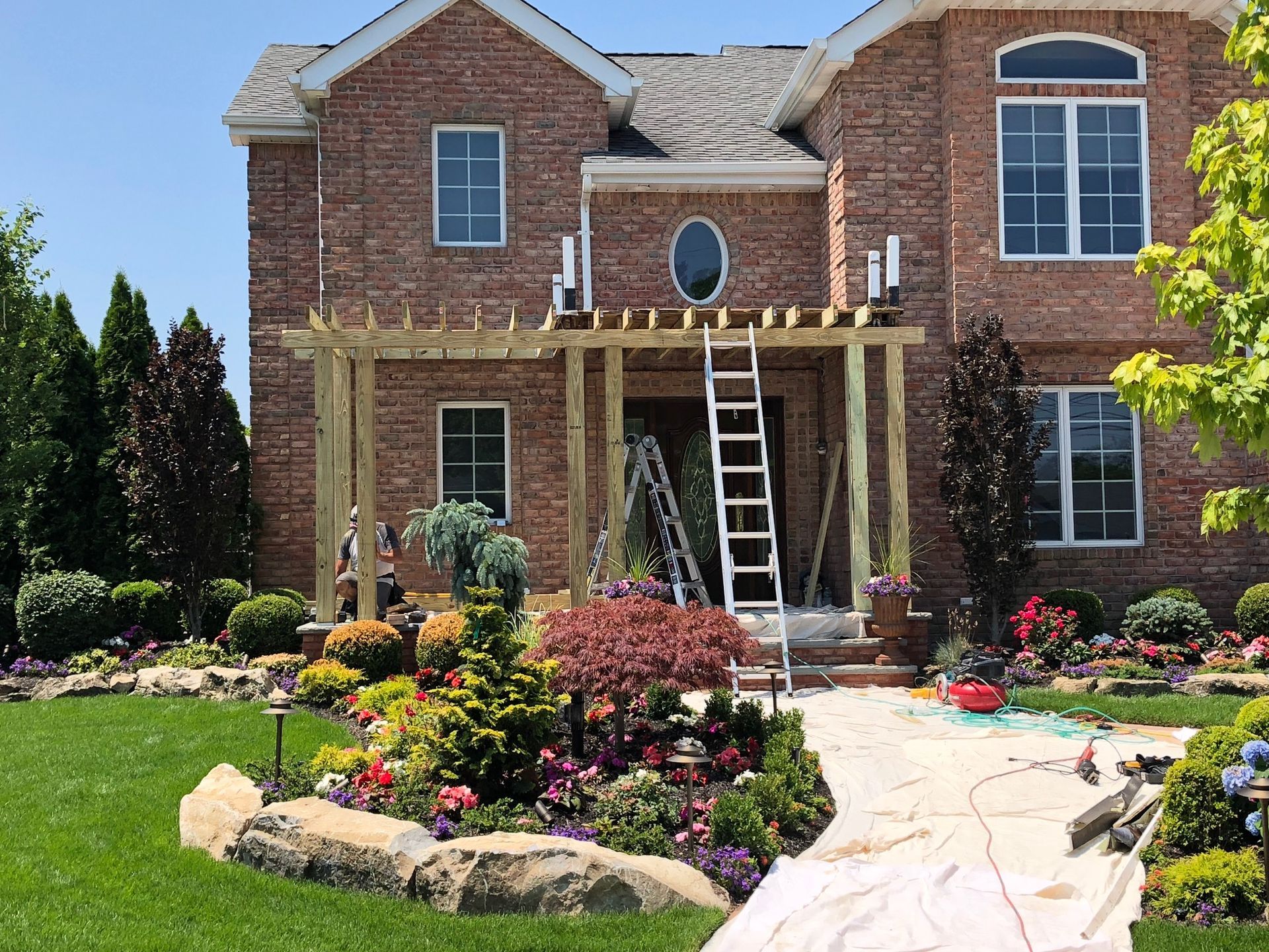 A brick house with a partially constructed wooden pergola over the front entrance, featuring a ladder and landscaping.
