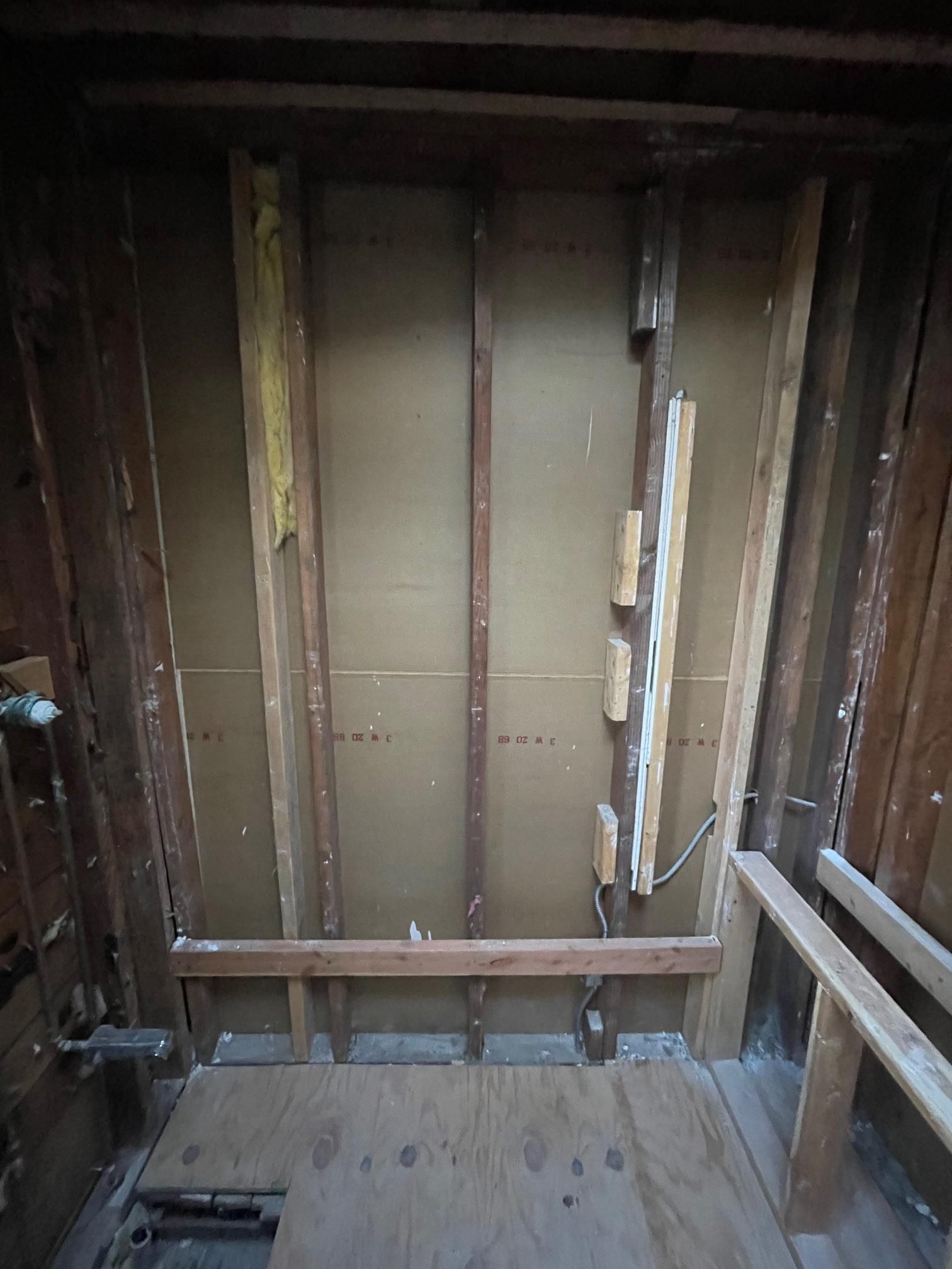 An interior wall frame during a remodel, showing exposed wooden studs, a plywood subfloor, and rough plumbing.