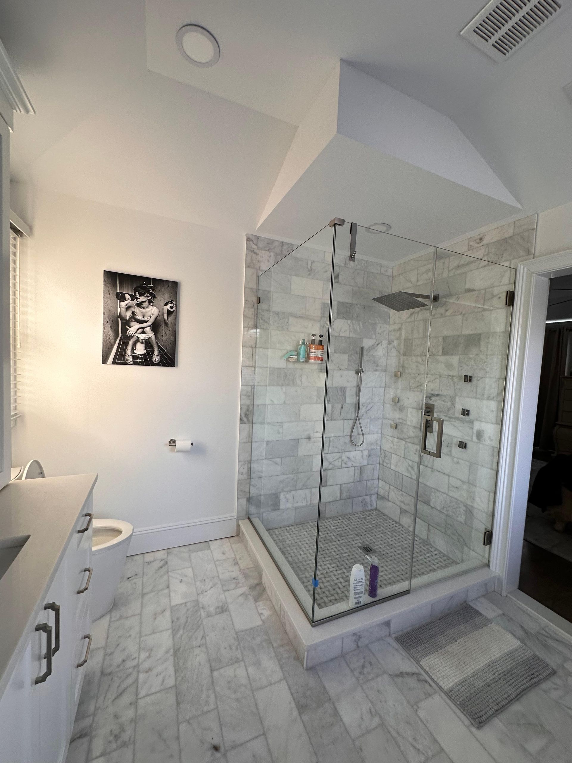 A bathroom with a white marble floor, a glass-enclosed stone shower, a white vanity, and black-and-white wall art.