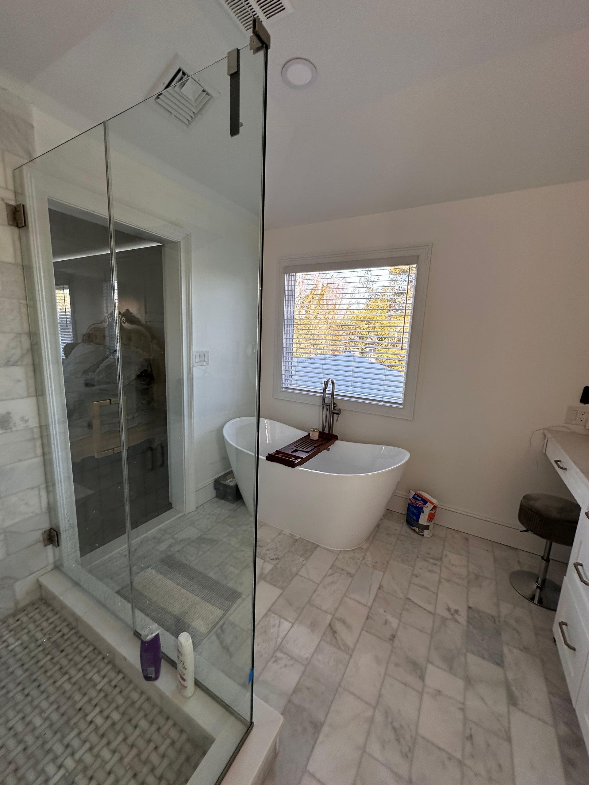 A bright, modern bathroom featuring a white soaking tub by a window and a large glass-enclosed shower on marble floors.