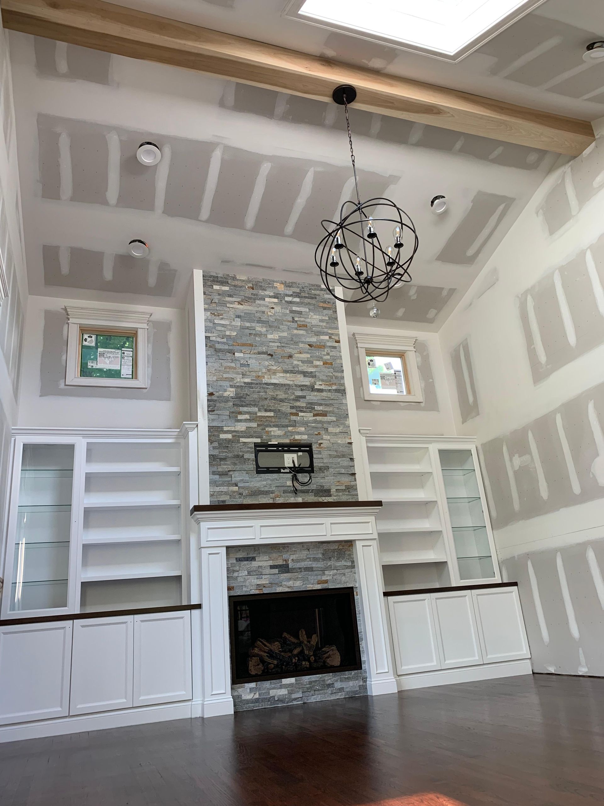 A stone fireplace with white built-in shelves in a room under construction with unfinished drywall and a chandelier.