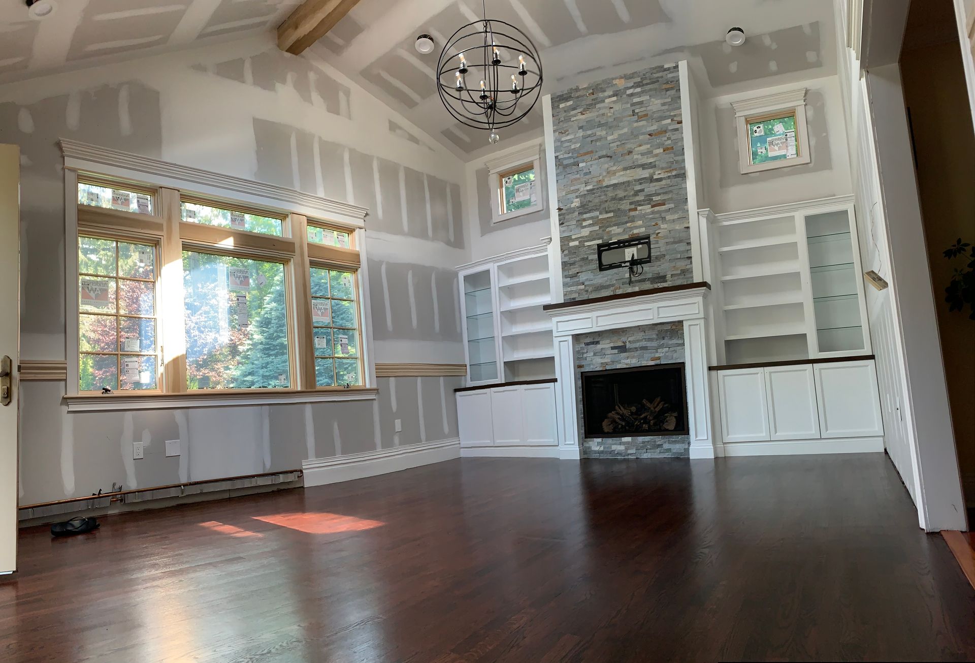 A room with dark wood floors, unfinished drywall walls, a stone fireplace, built-in shelves, and a large window.