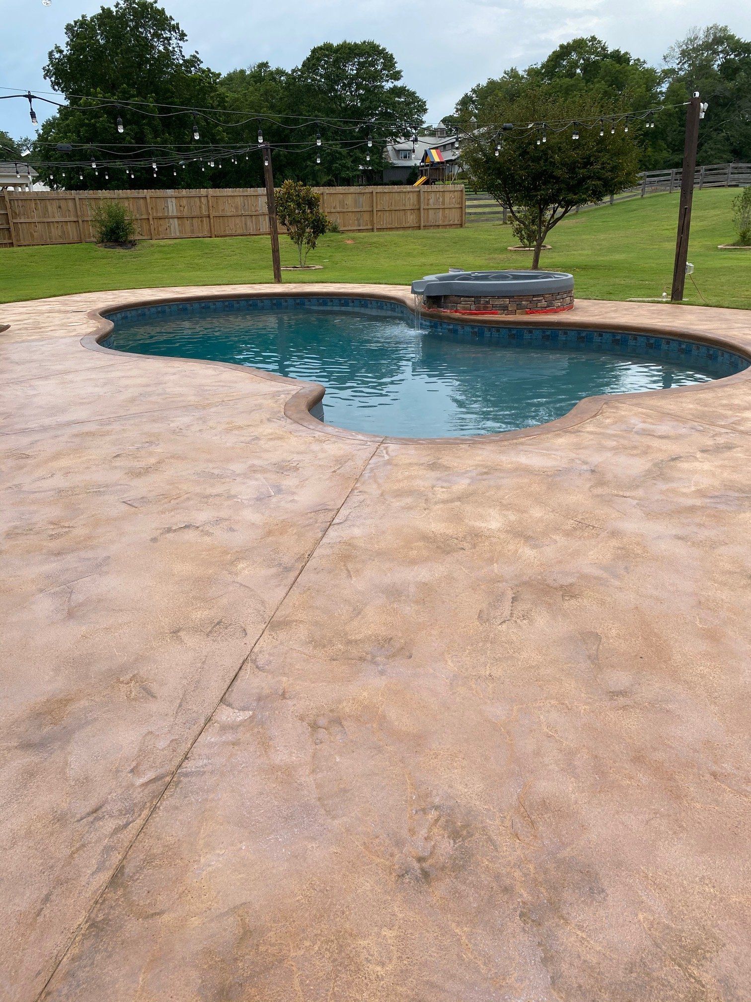 Backyard pool with blue water, curved concrete deck, and grassy yard with trees and a fence.