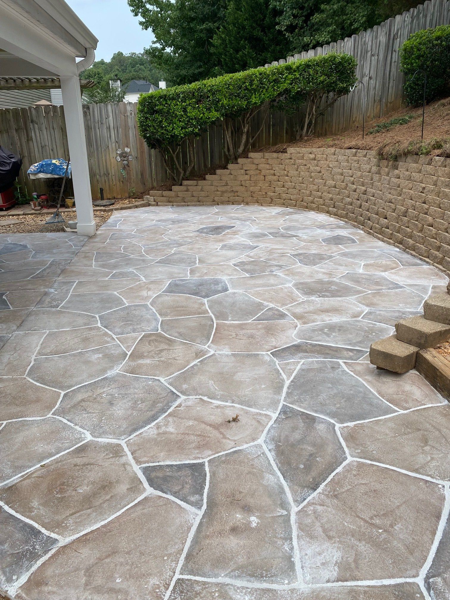 Stone patio in a backyard beside a house, with a fenced, sloped landscape and greenery in the background