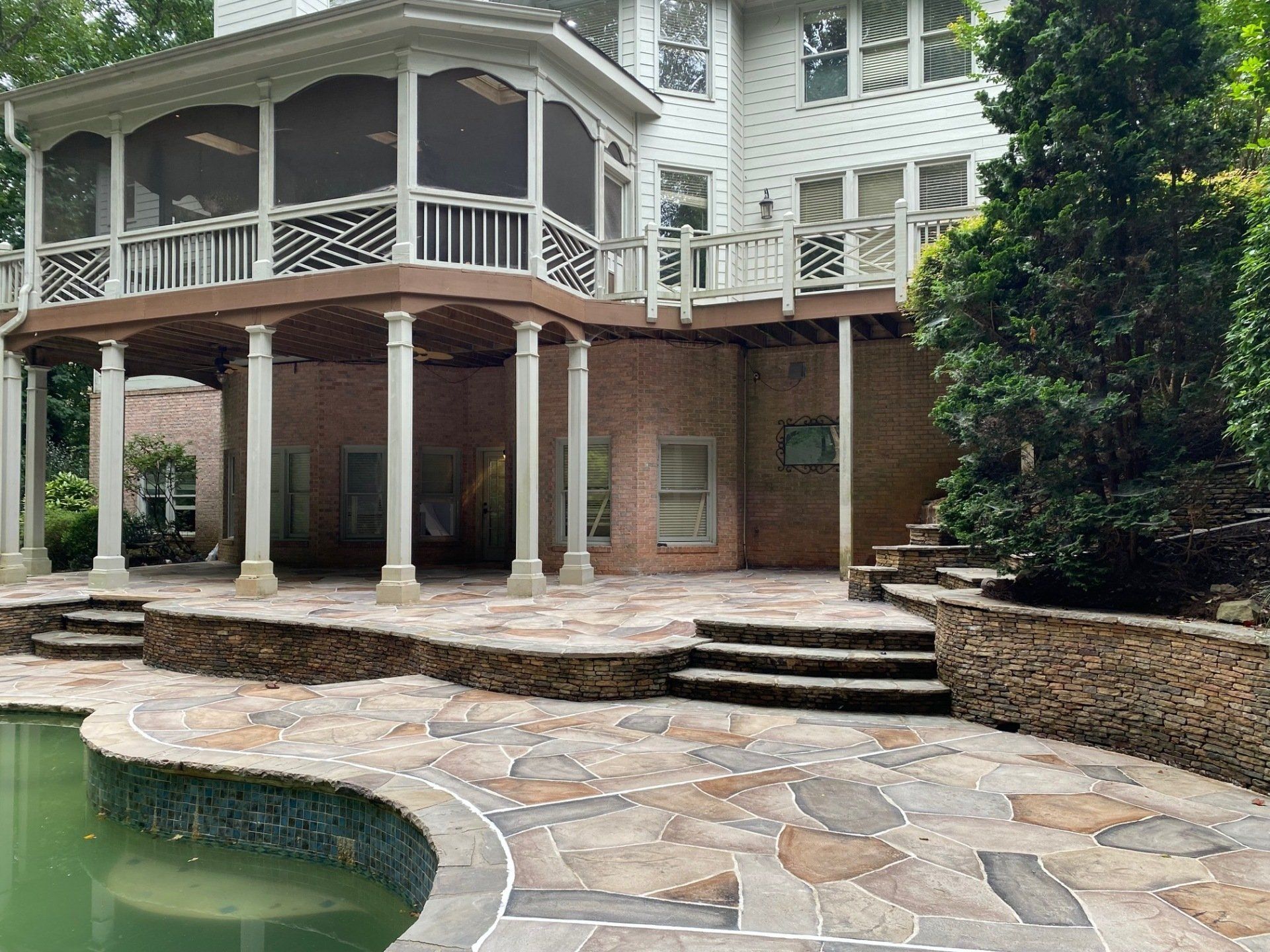 Large white house with wraparound porch, stone patio, steps, and a curved pool in front