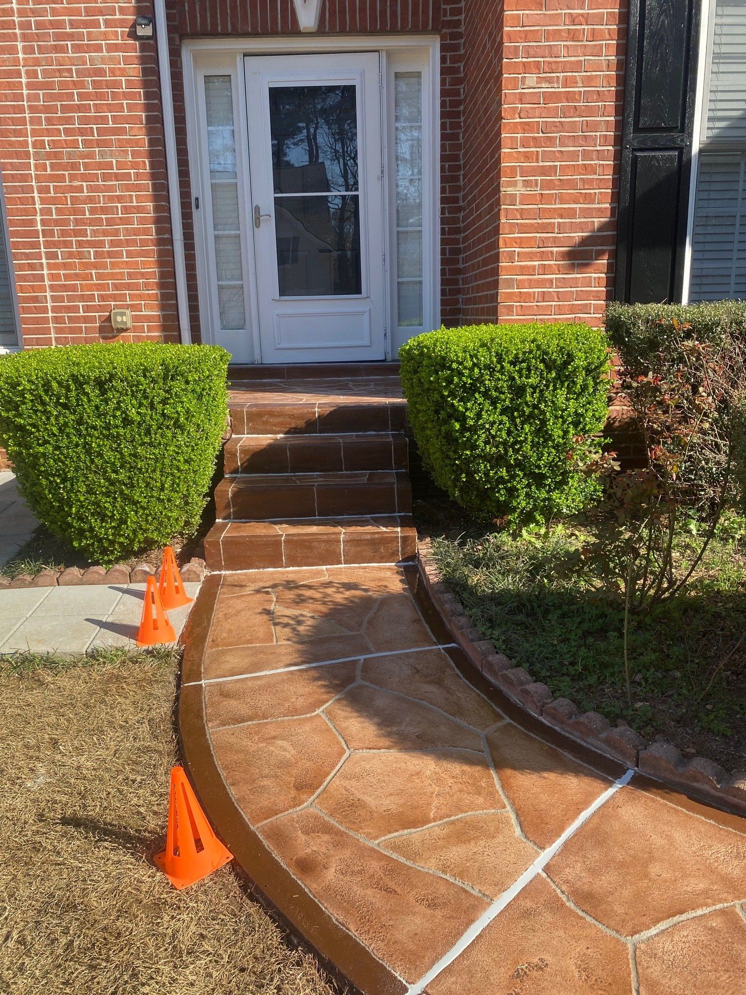 Brick front entry with steps, white door, trimmed bushes, and orange cones marking a curved walkway