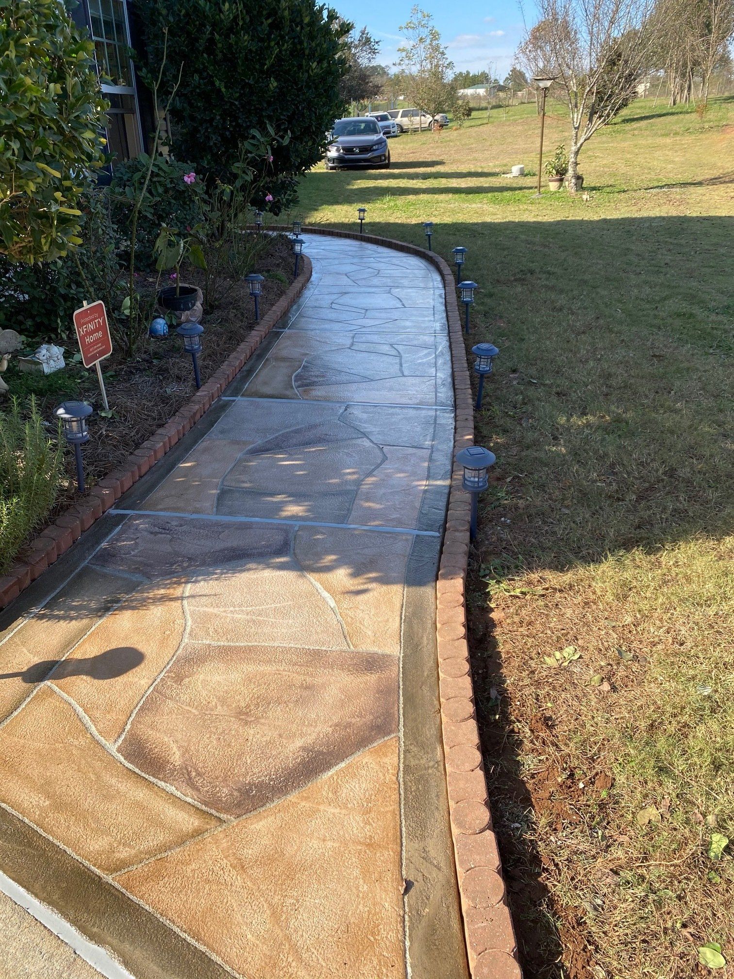 Curved concrete walkway with wet patches beside a grassy lawn and shrubs, leading to a parked car.