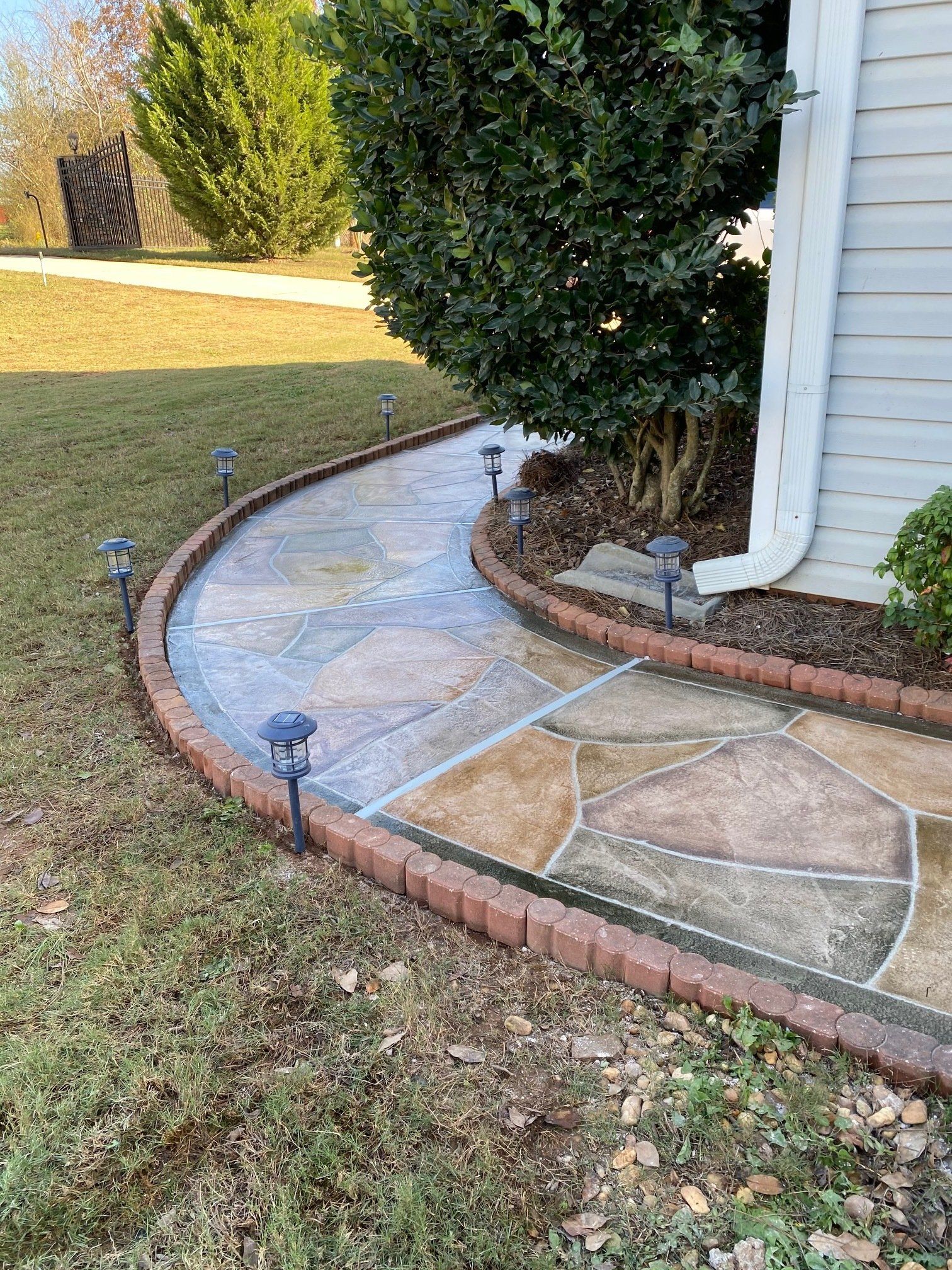 Curved stone garden walkway with brick edging, solar lights, and a tree beside a house