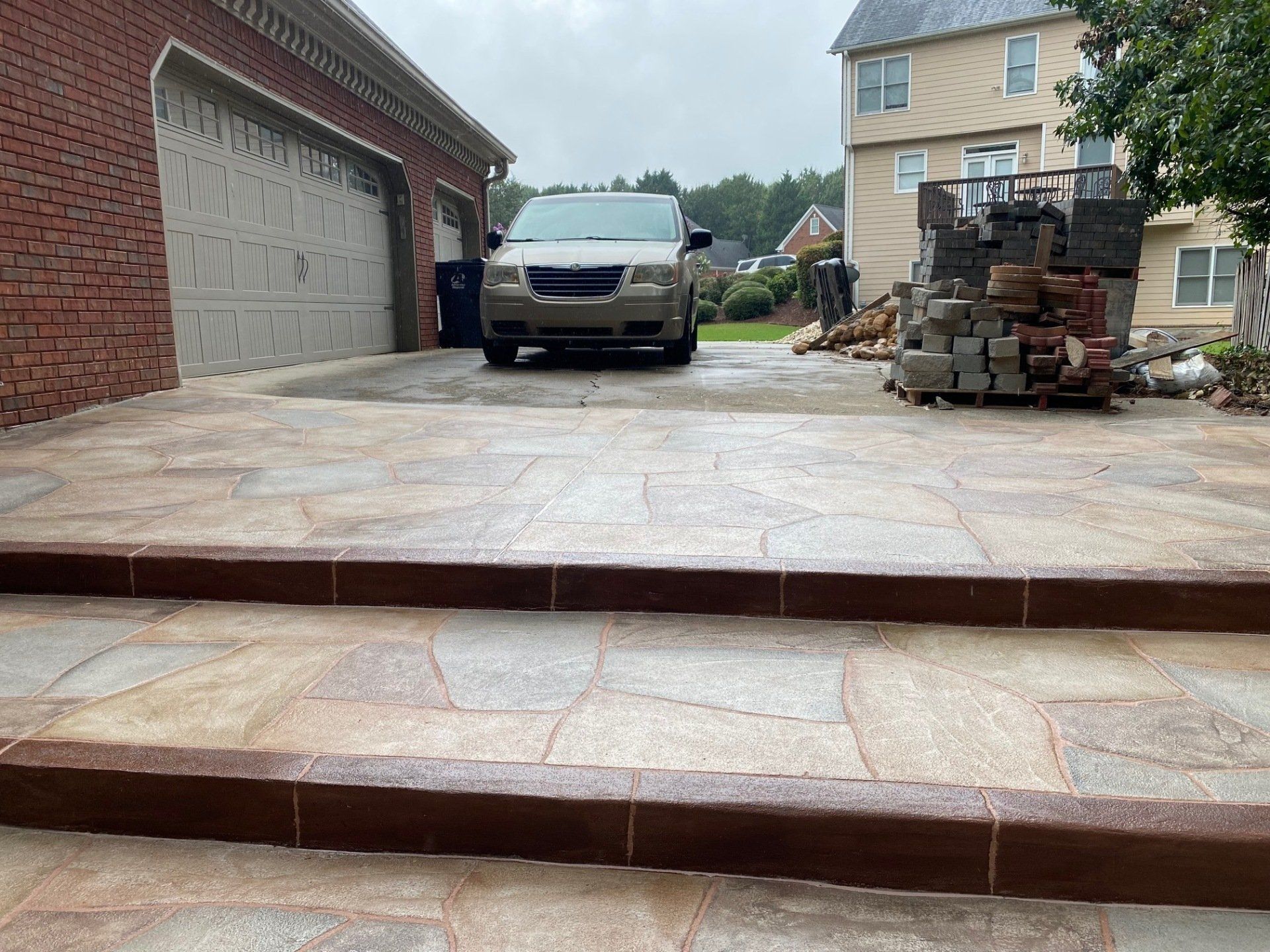 Brick-paver driveway with a black SUV, garage on the left, and stacked bricks beside a house.