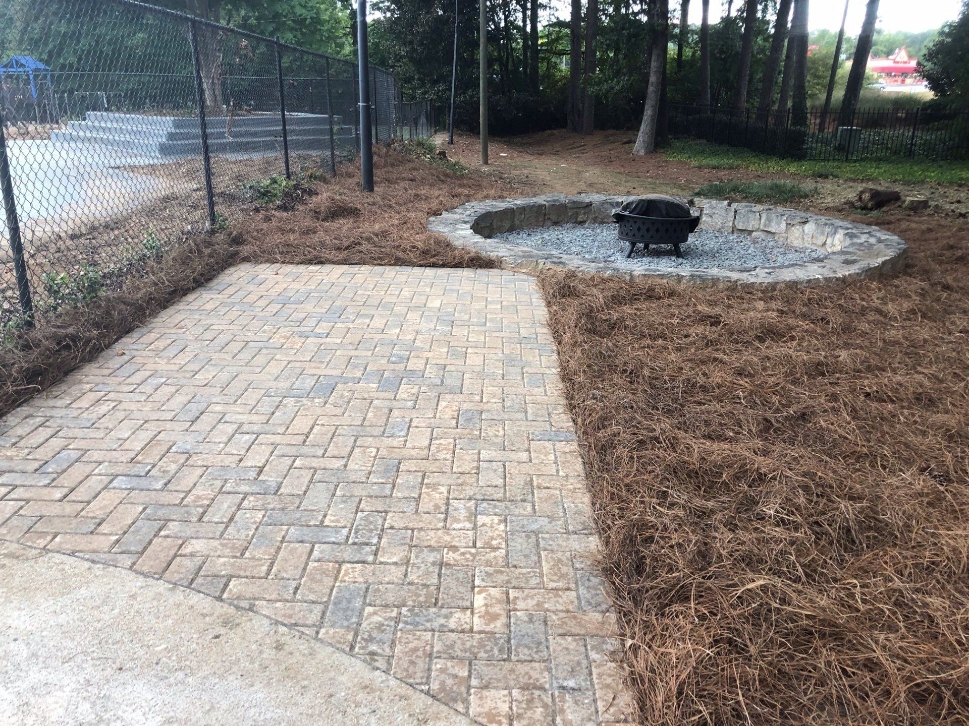 Brick walkway beside a fenced wooded area and a circular gravel fire pit surrounded by mulch.